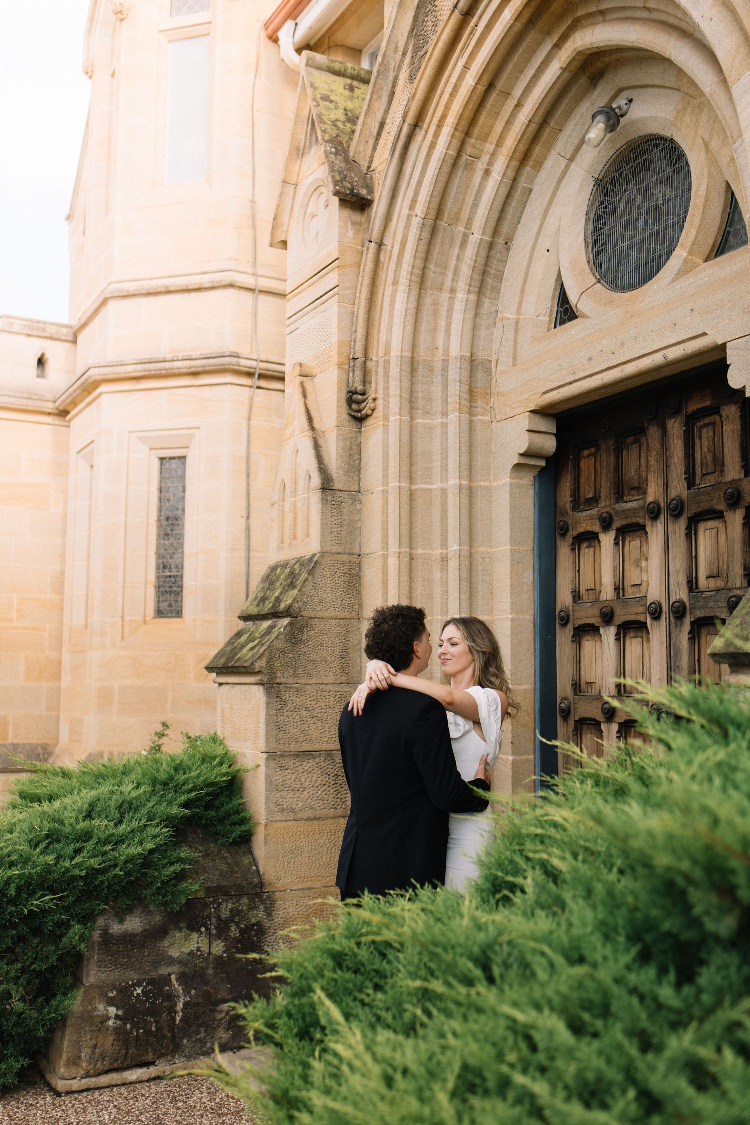 Bride and groom embracing outside a historic stone church with ornate wooden doors and lush green shrubs.