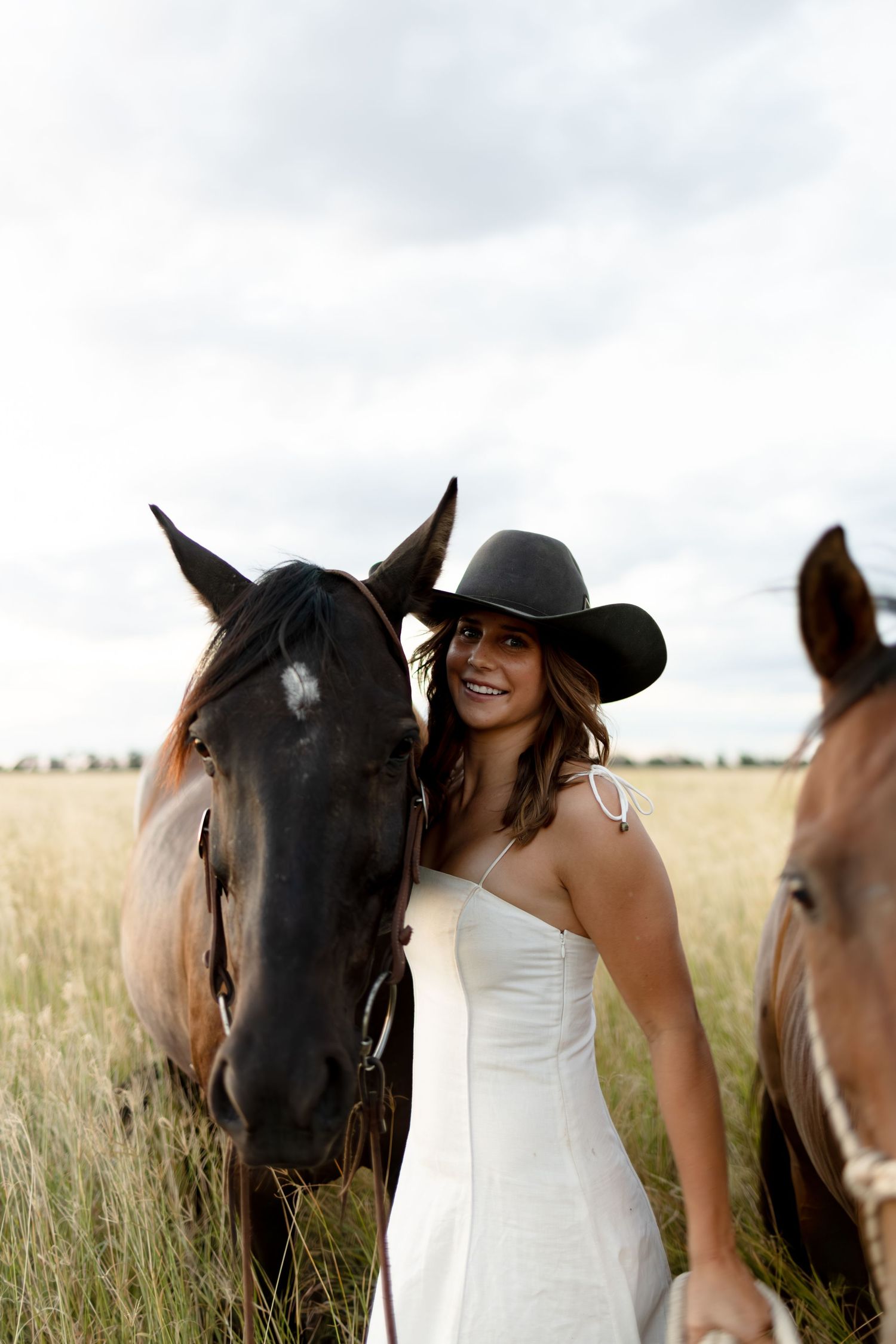 Smiling woman in white dress and black cowboy hat stands close to a dark brown horse in a field.