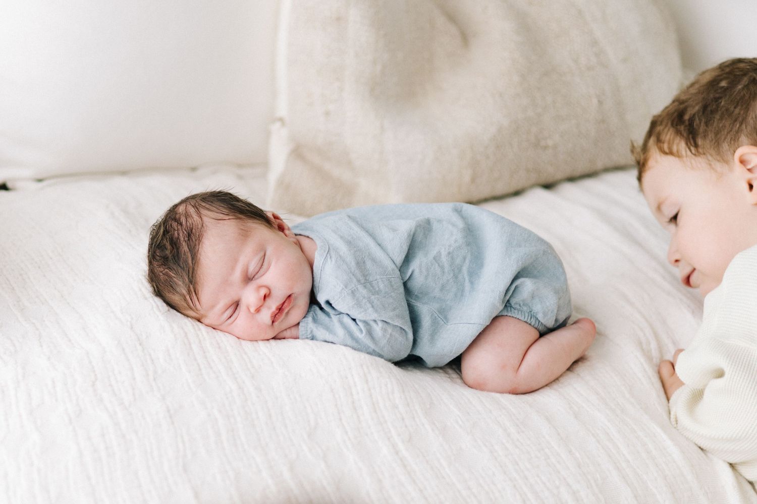 Newborn baby in blue romper sleeping on white bed while older sibling watches lovingly nearby.