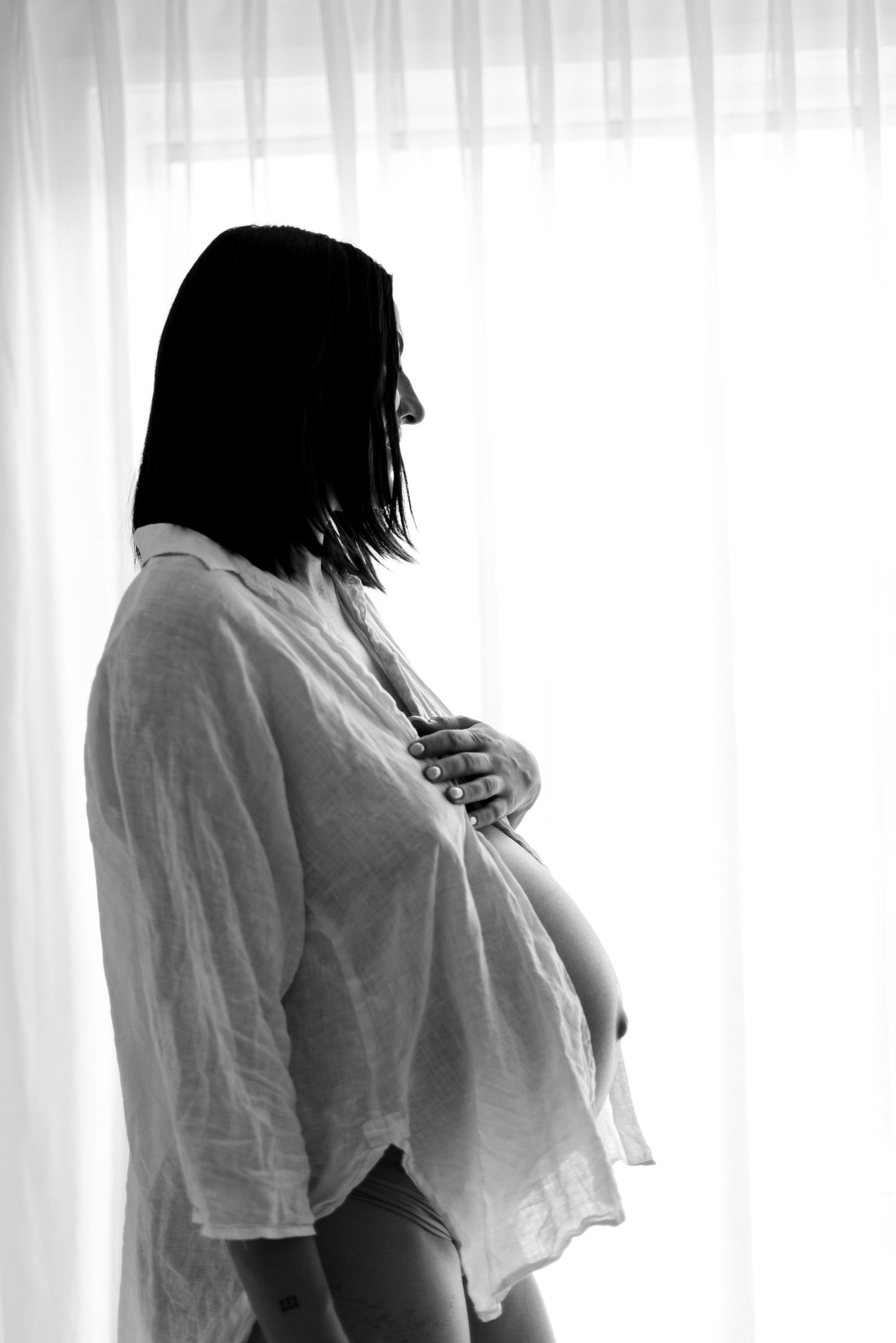 Black and white silhouette of pregnant woman in open white shirt cradling bump by window.