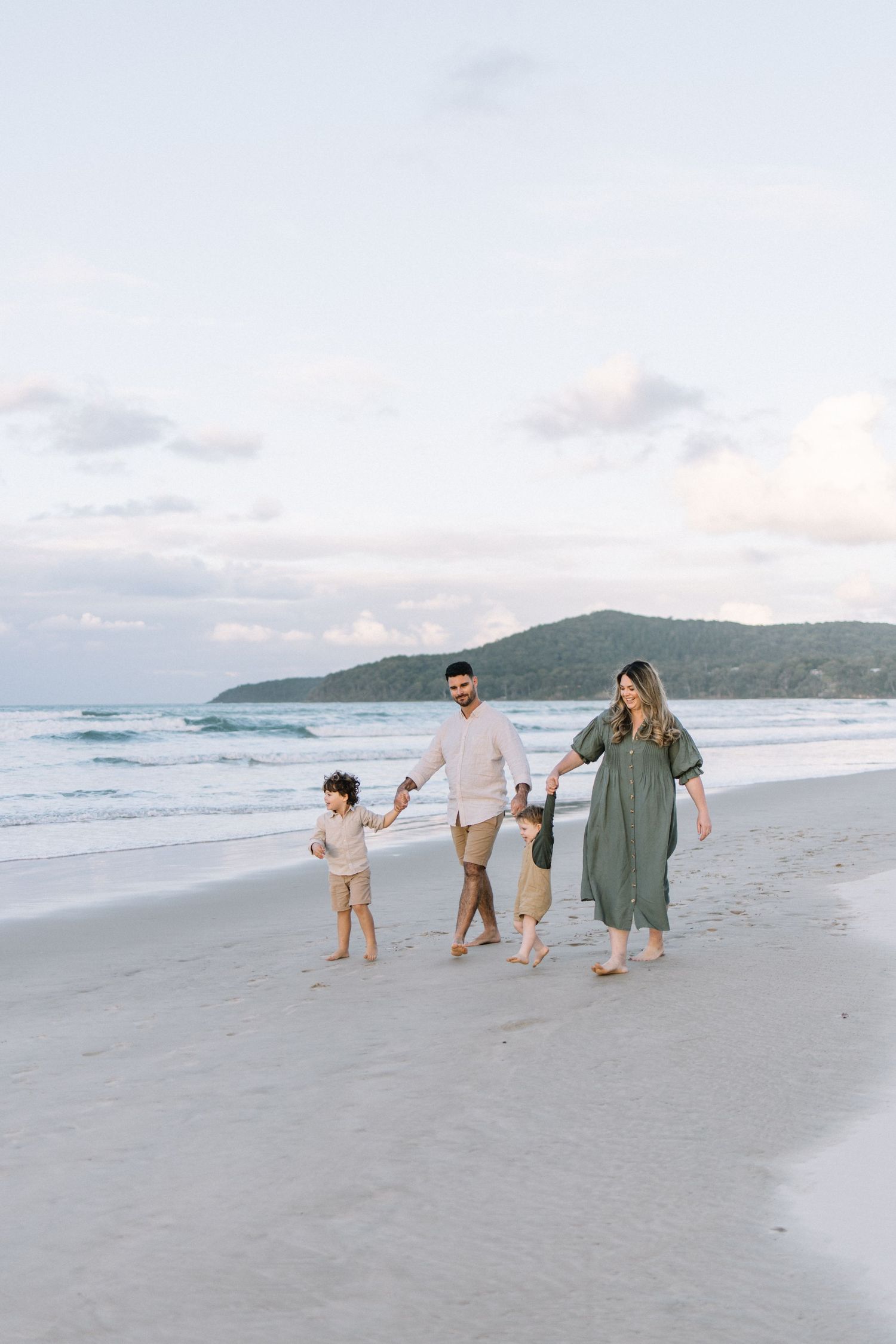 Family of four walking hand-in-hand on a sandy beach at dusk, with ocean waves and green hills in the background.
