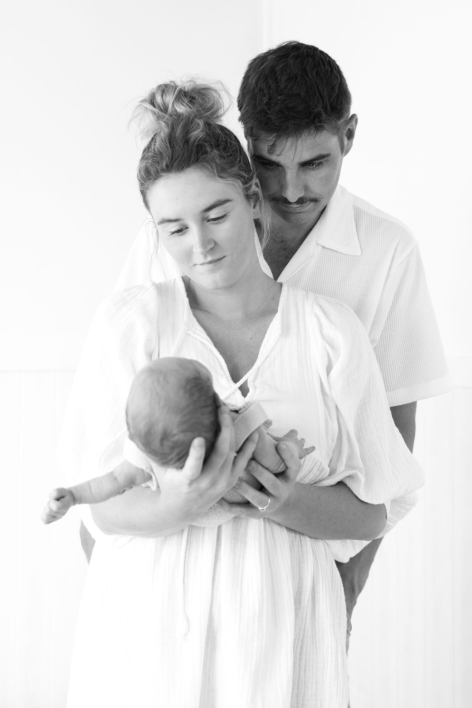 Black and white portrait of new parents in white outfits lovingly holding their newborn.