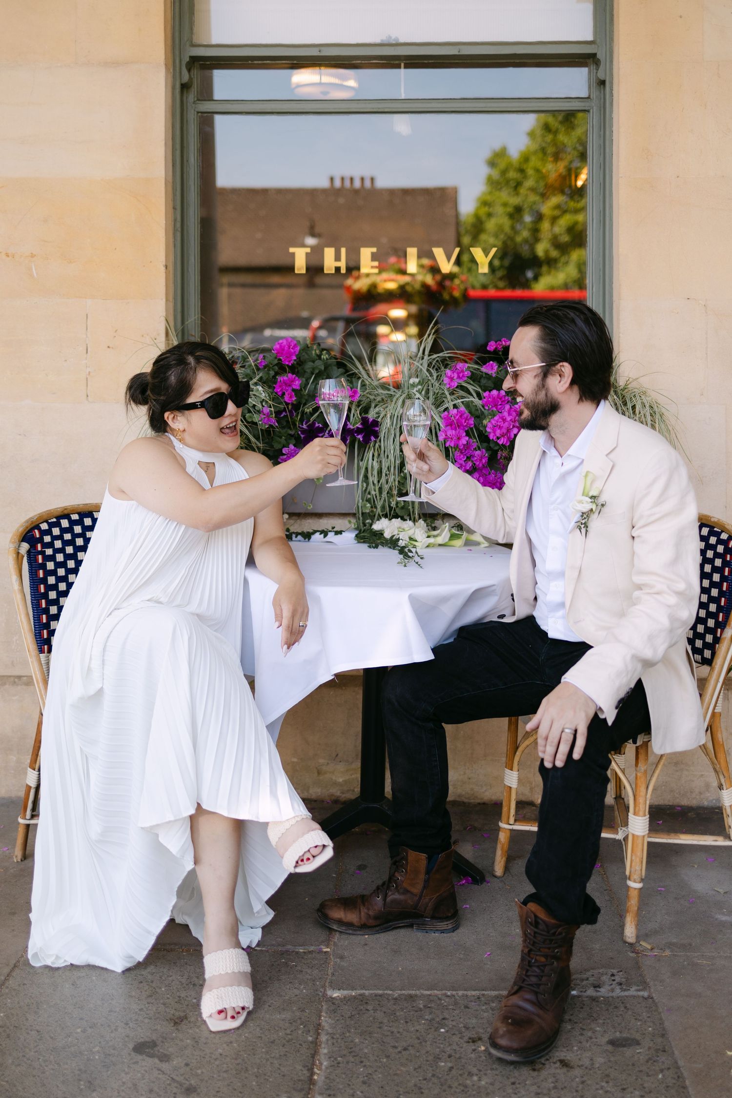 Bride and groom toasting with wine glasses at outdoor table outside The Ivy restaurant on their wedding day.