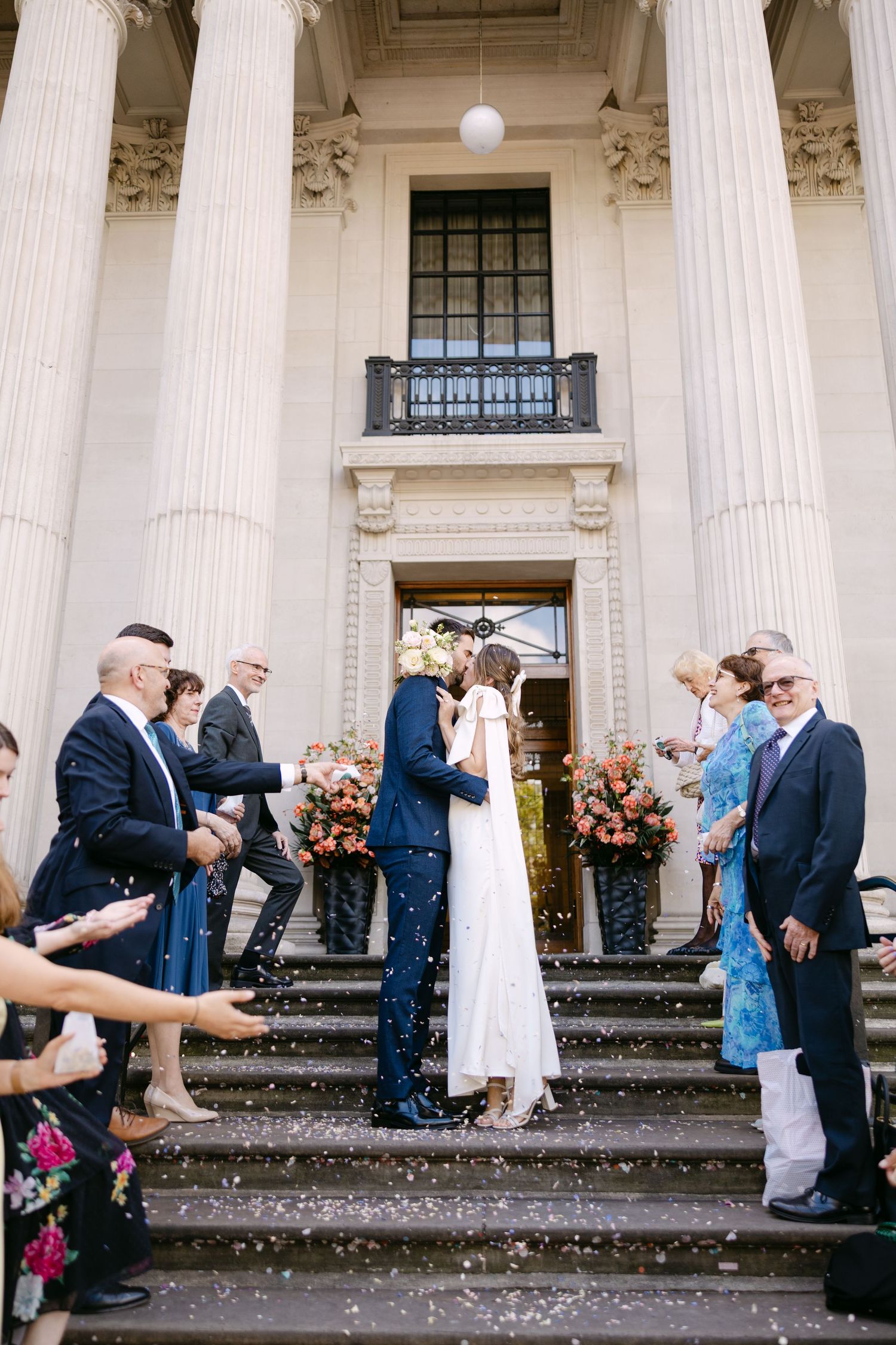 A wedding ceremony on grand stone steps with white columns and gathered guests throwing confetti at The Old Marylebone Town Hall.