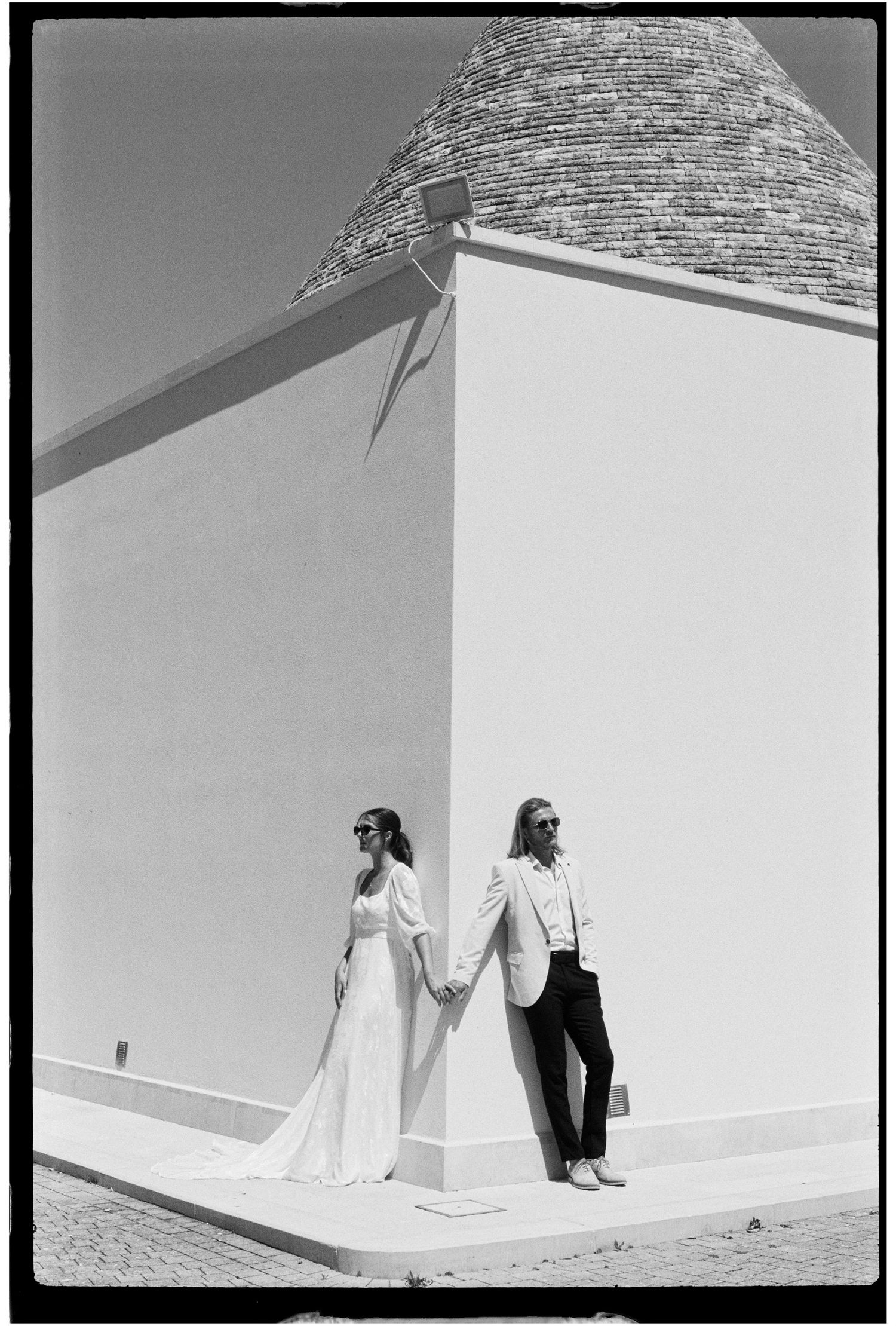 Black and white photo of a bride and groom holding hands against a white trullo building wall in Alberobello, Italy.