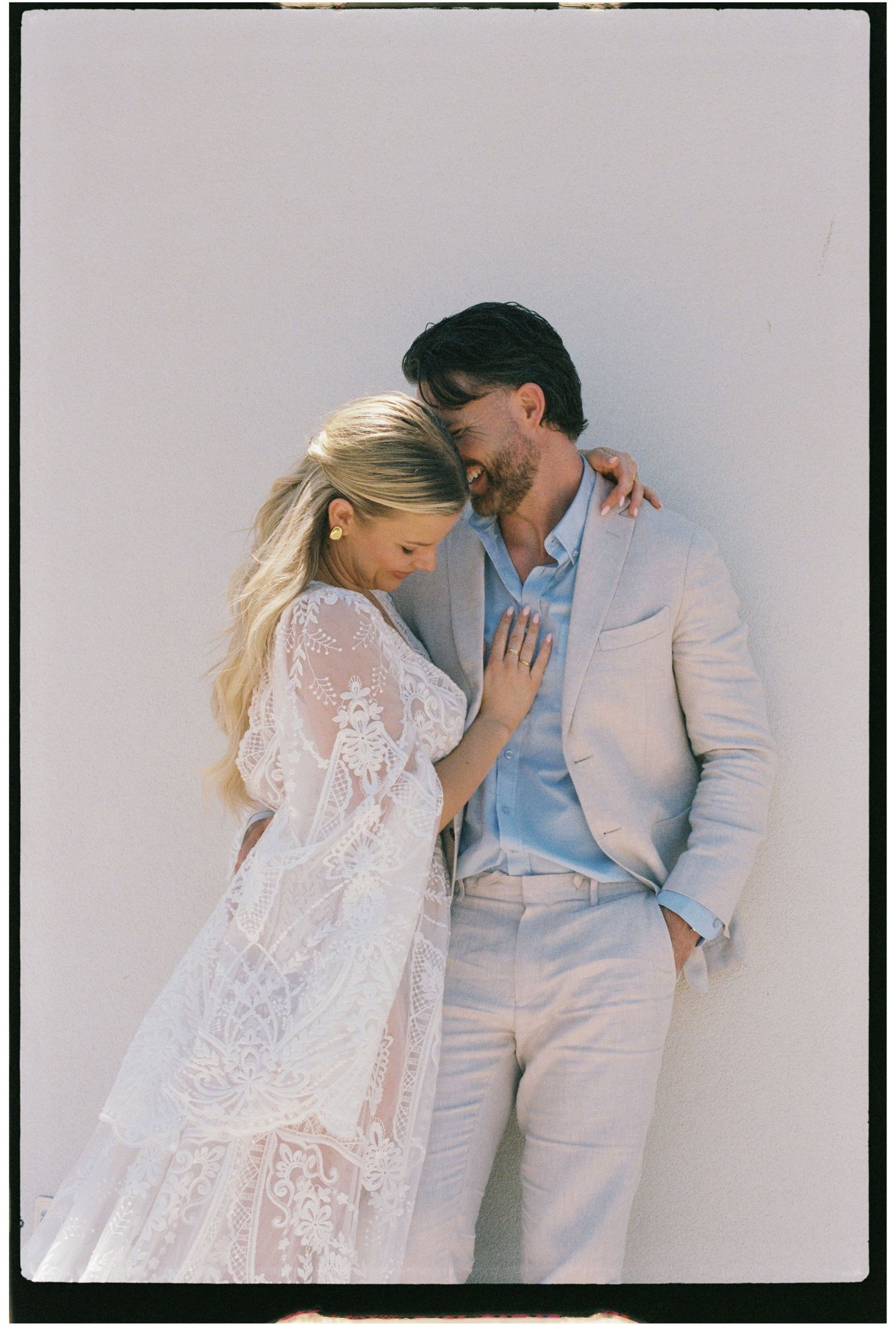 Couple in wedding attire share a tender embrace leaning against a bright white wall outdoors.