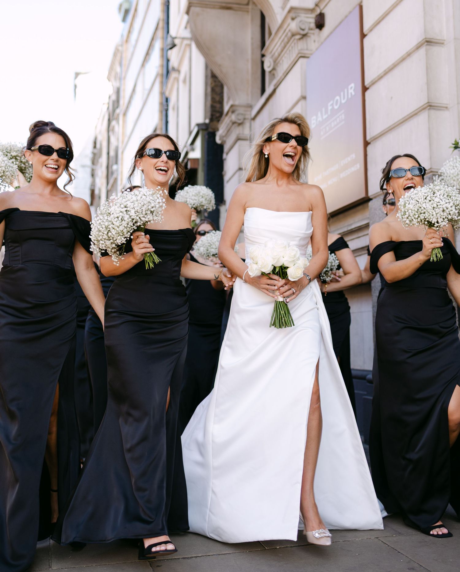 Bridesmaids in elegant black dresses pose with a bride in white holding bouquets against a light building exterior.