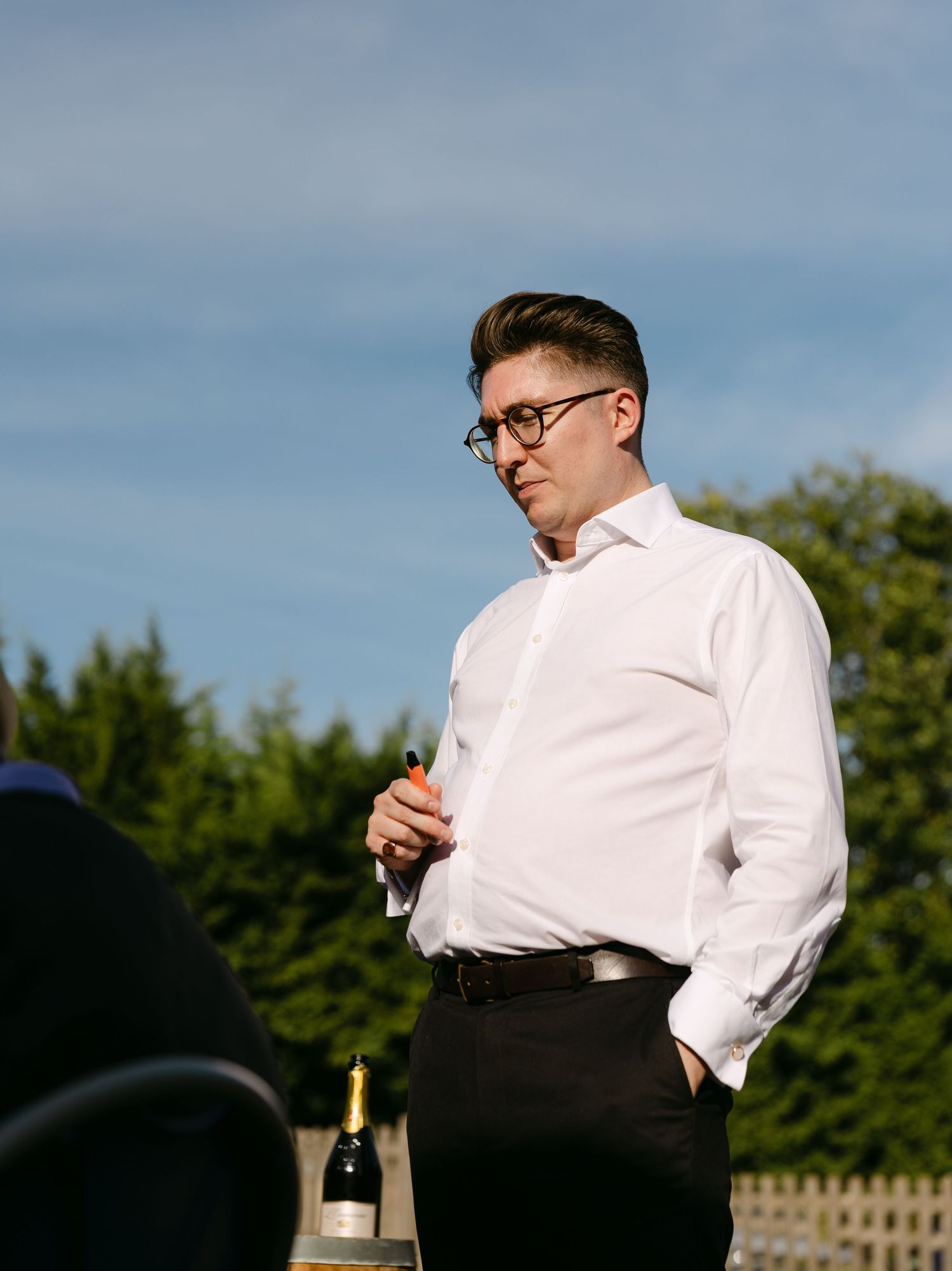 A person in formal attire stands outdoors against a blue sky backdrop.