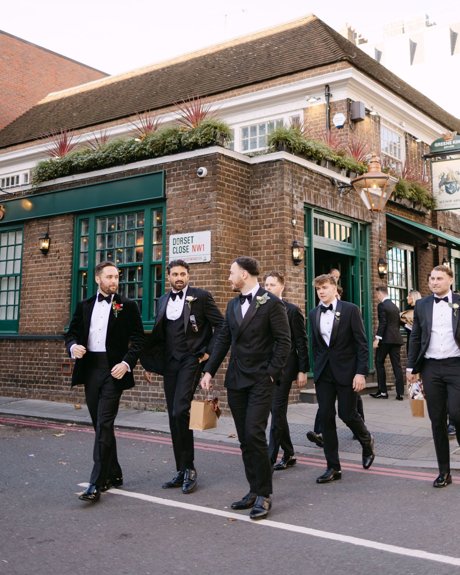 Wedding party walks down street in formal attire past brick building with green storefront.