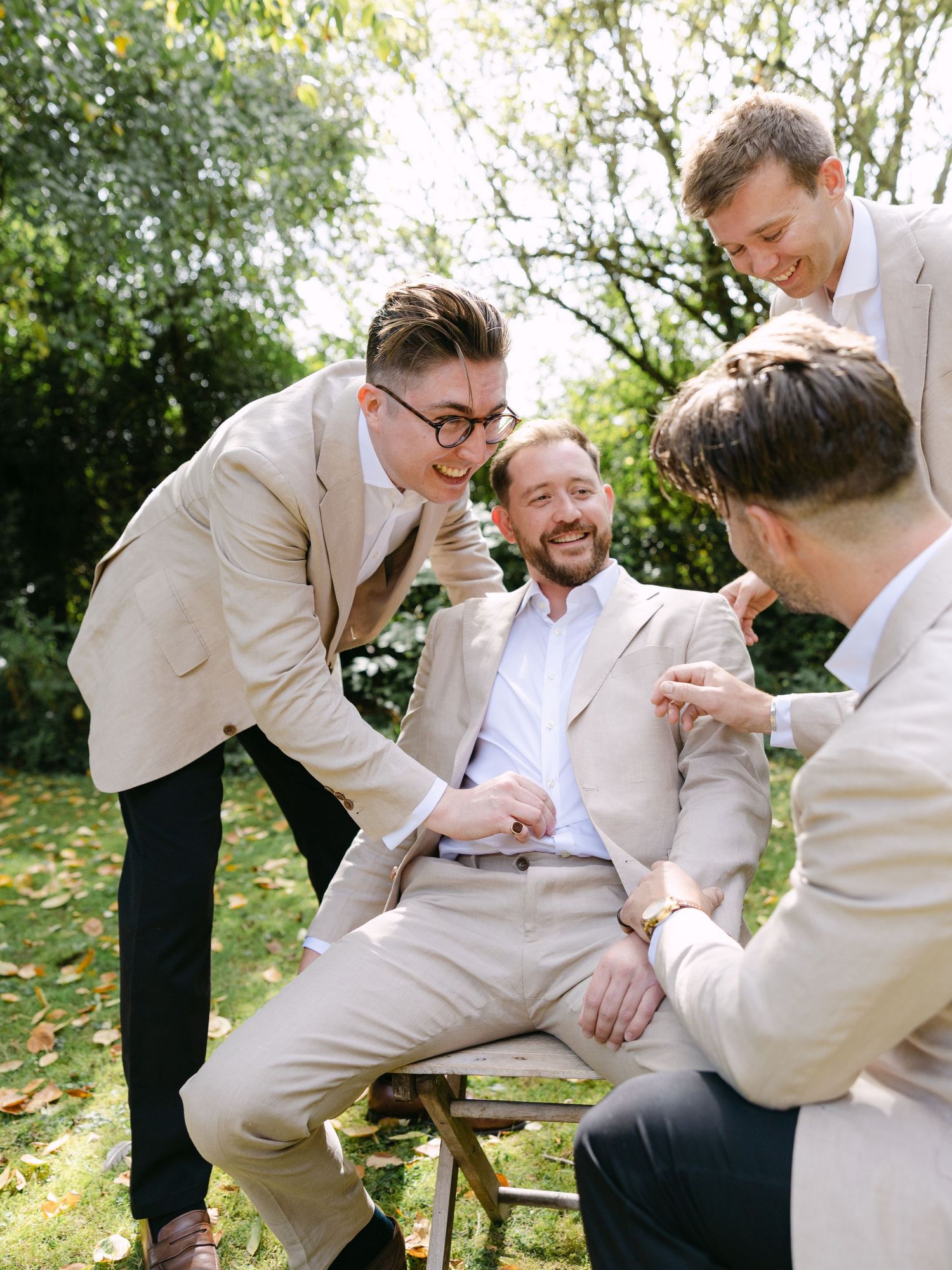 Wedding party members in light colored suits sharing laughs on outdoor bench.