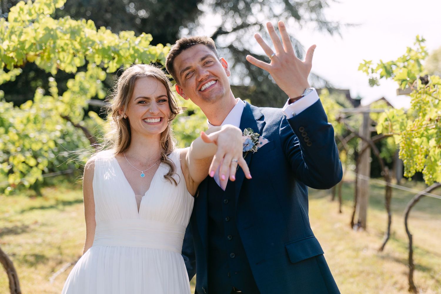 Smiling newlyweds show off wedding rings outdoors at vineyard, bride in white dress, groom in navy suit.