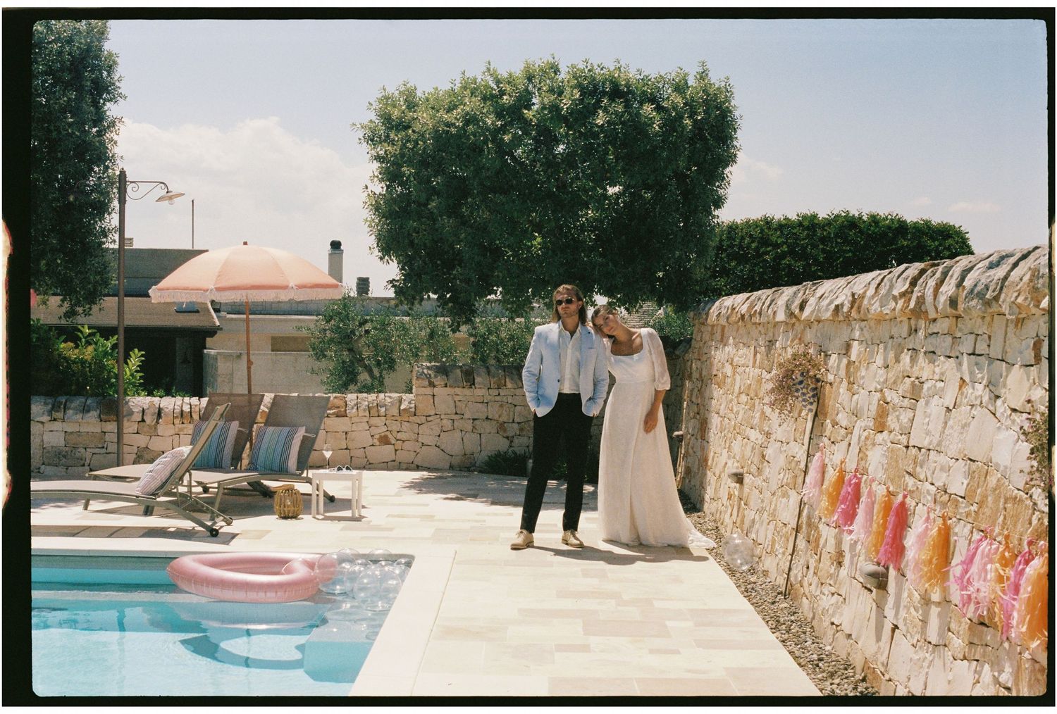 Couple standing by a pool next to a stone wall at a sunny Puglia trullo estate venue.
