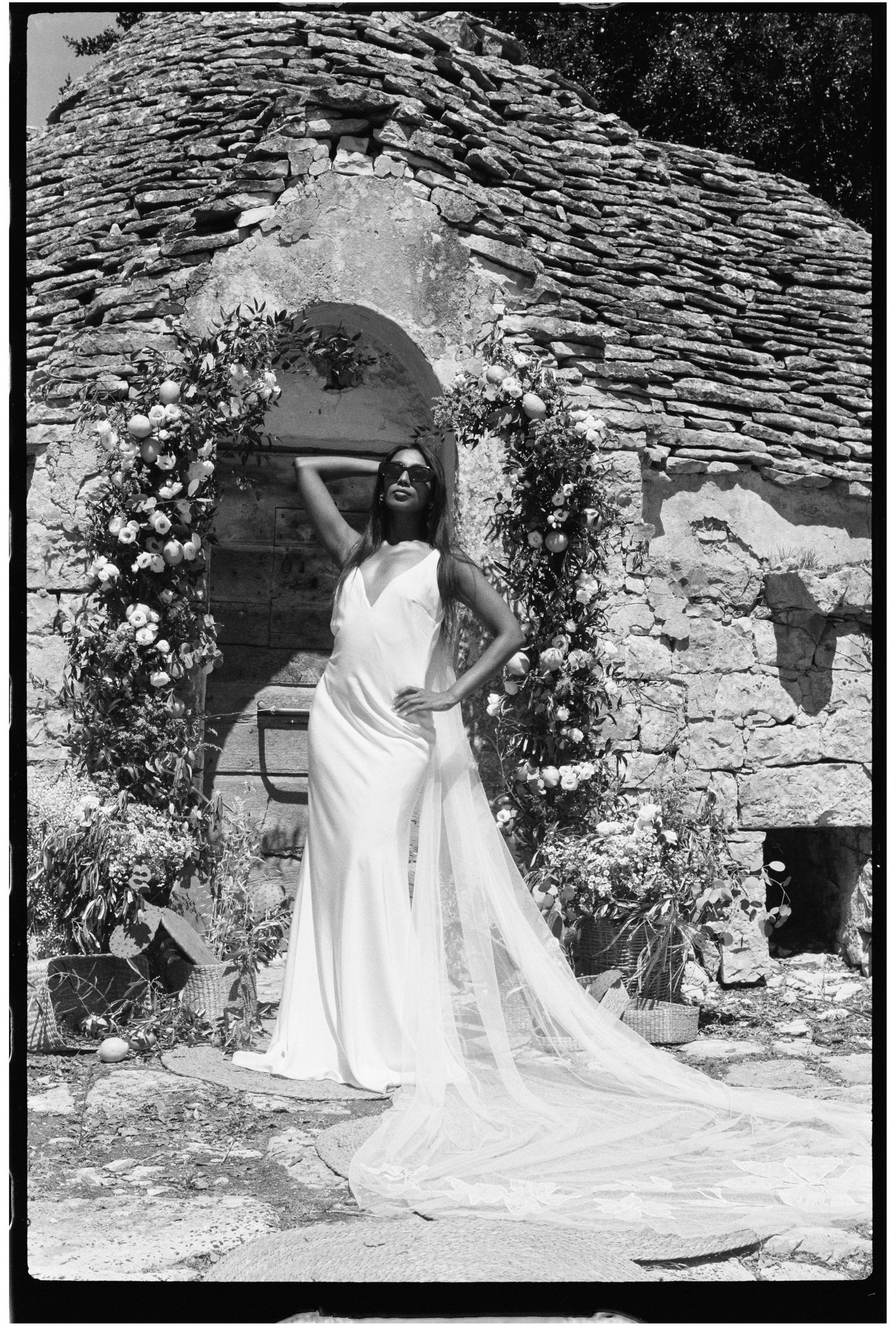 Bride in white gown poses before a floral arch at a rustic stone building on a sunny day.