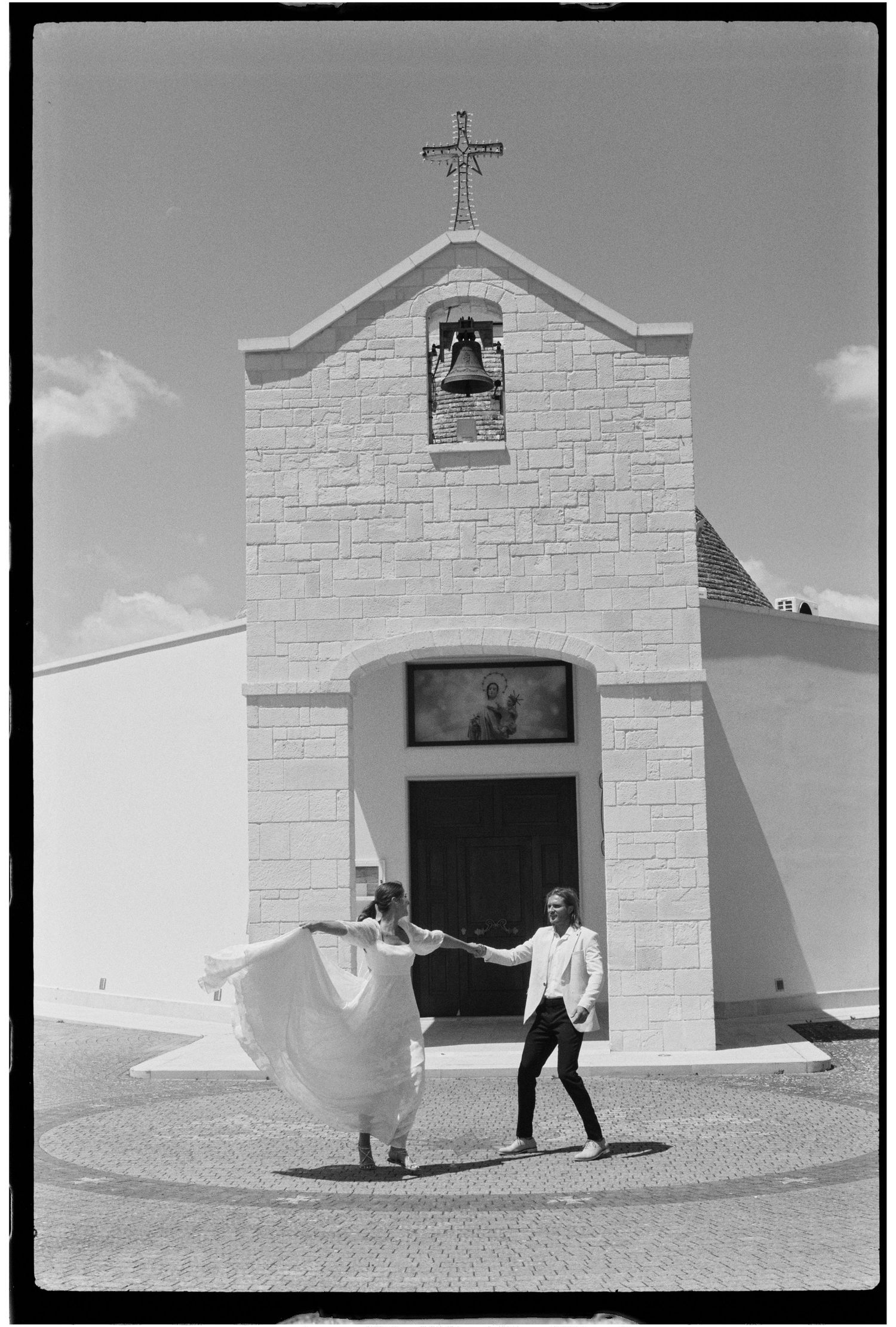 Black and white photo of a bride and groom dancing joyfully in front of a small white chapel.