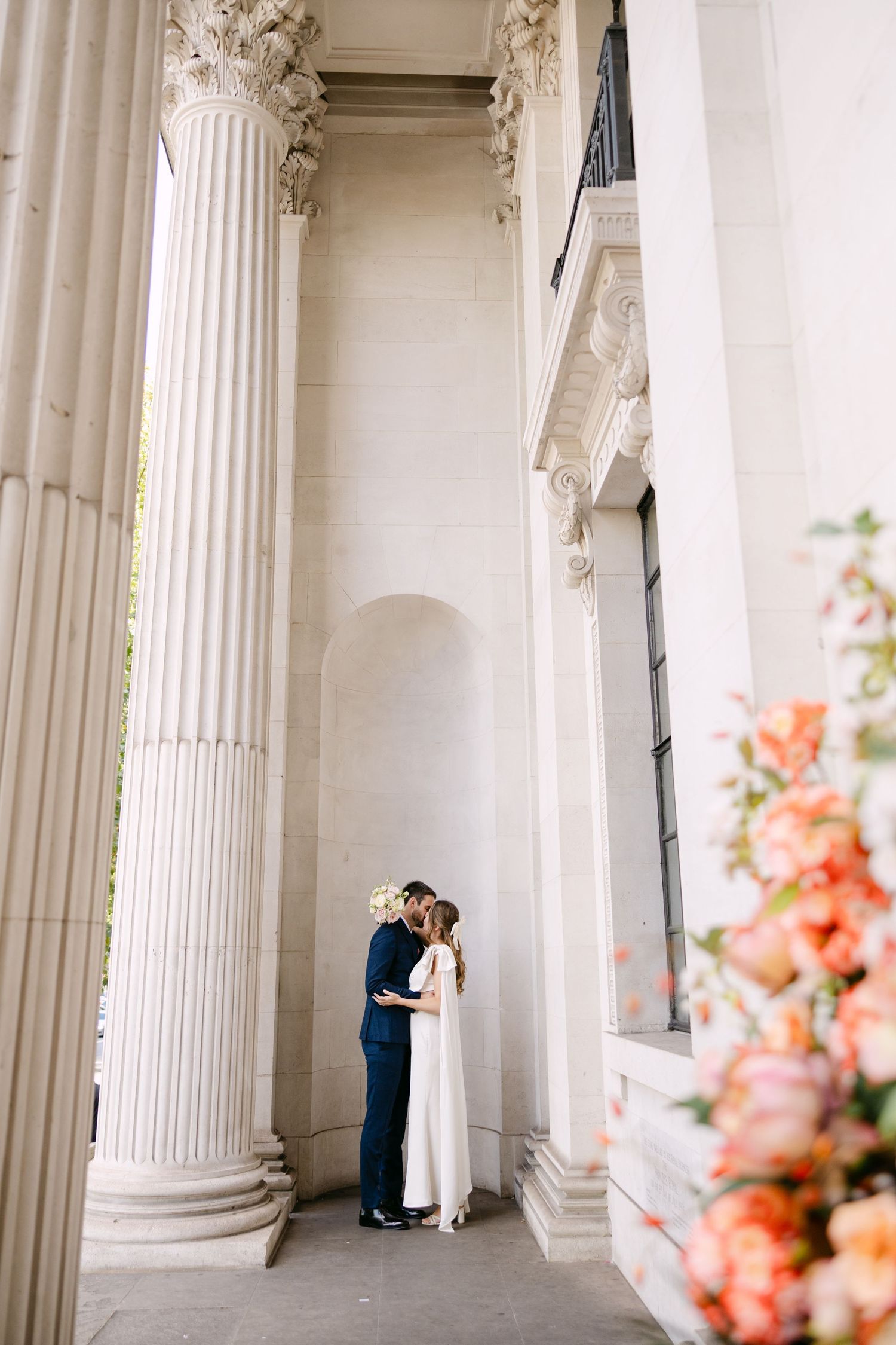 A couple shares an intimate moment between classical white columns with coral flowers in foreground at The Old Marylebone Town Hall.