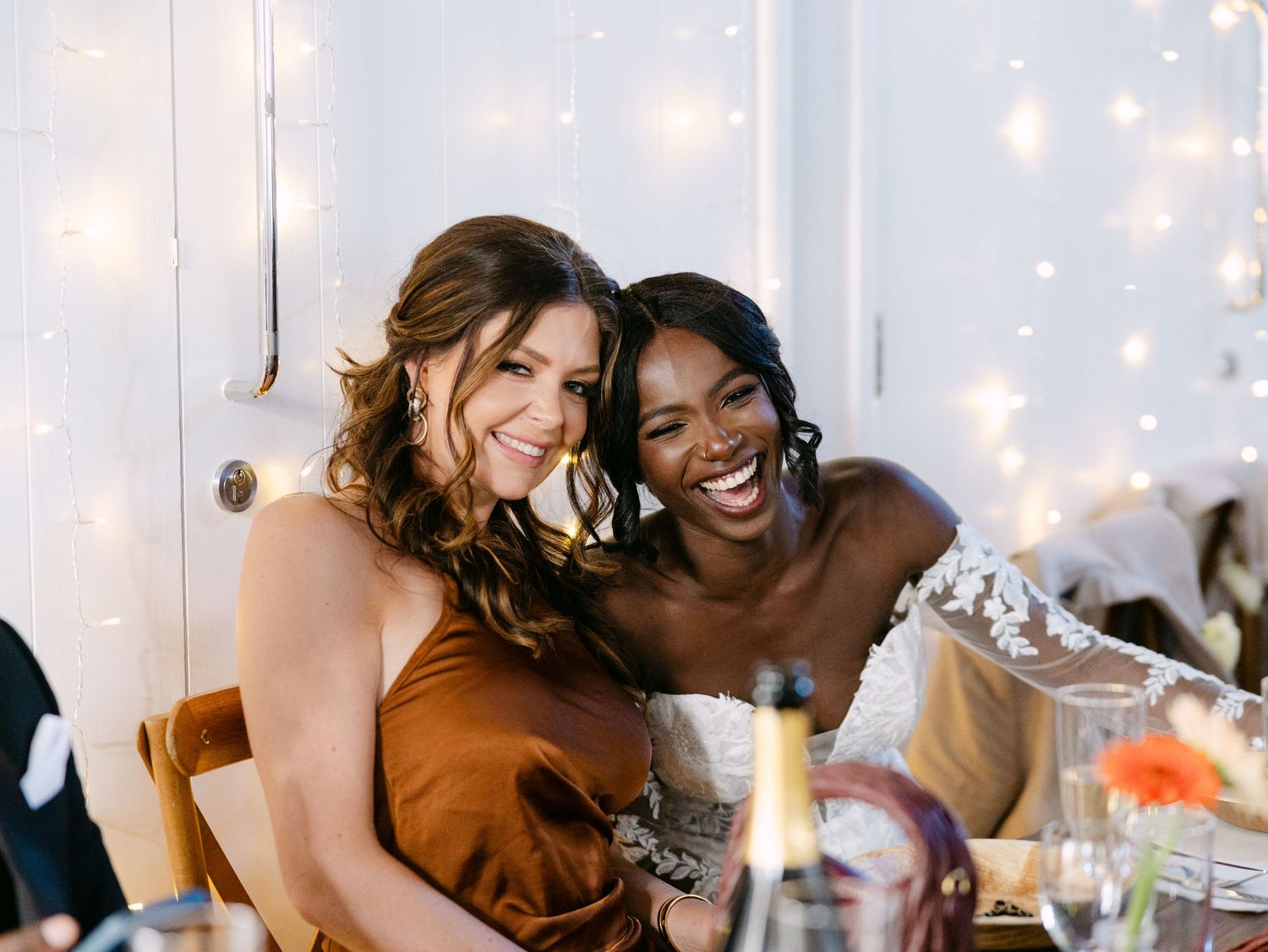 Two friends sharing a joyful moment at a celebration with string lights in the background.