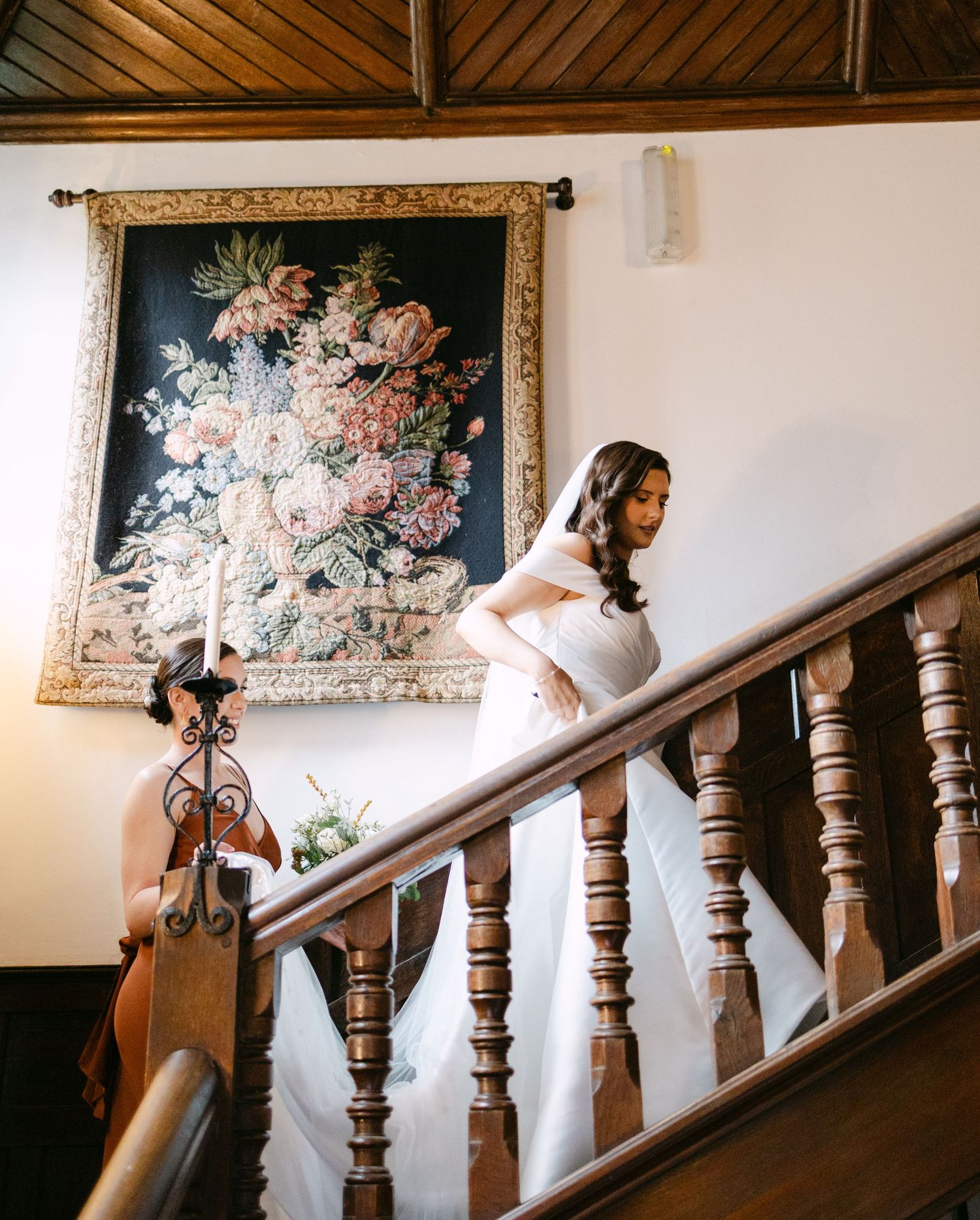 A bride in a white dress descends a grand wooden staircase with ornate floral artwork in the background.