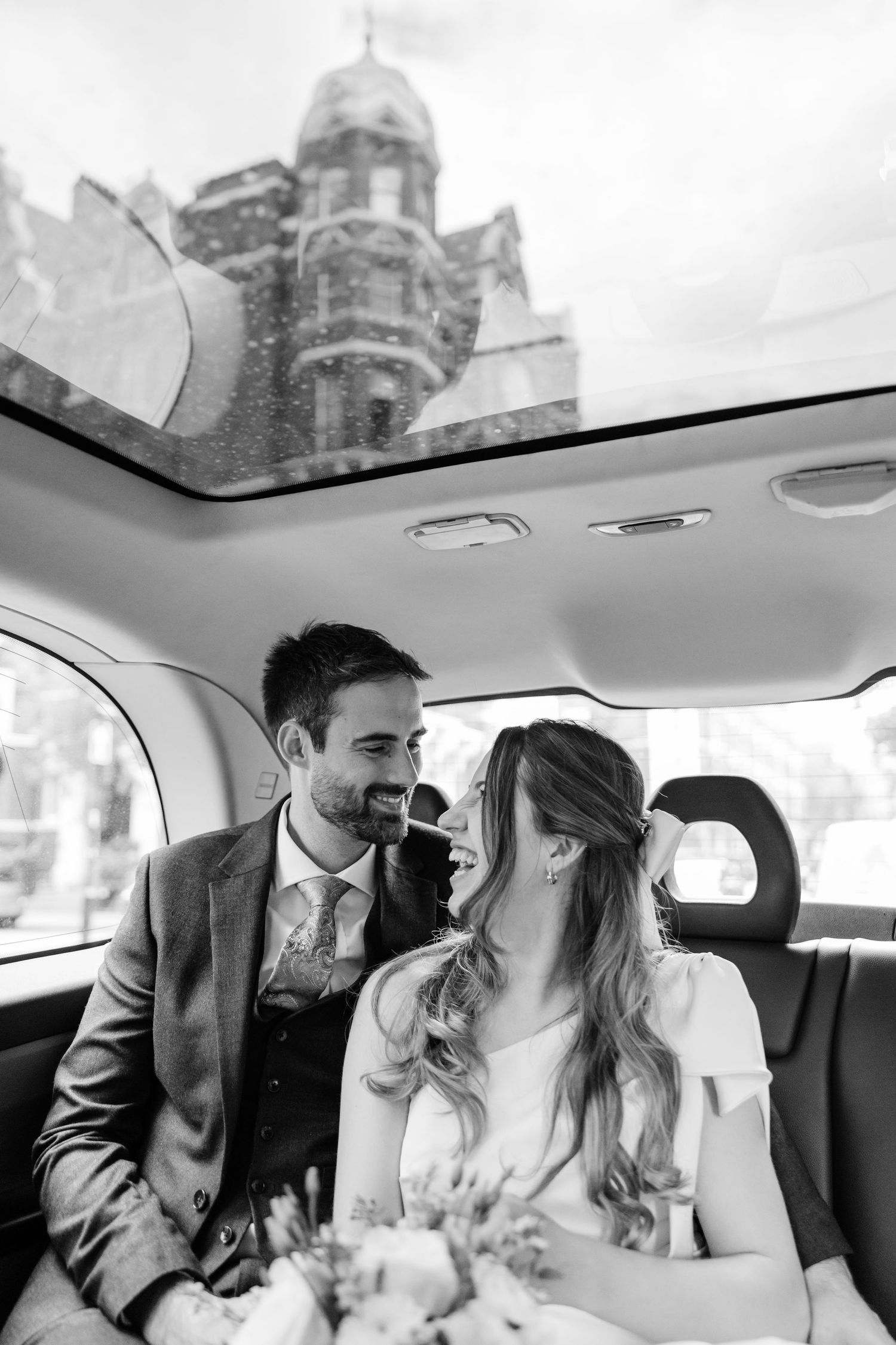 Bride and groom laughing in London taxi with glass roof showing city architecture black and white