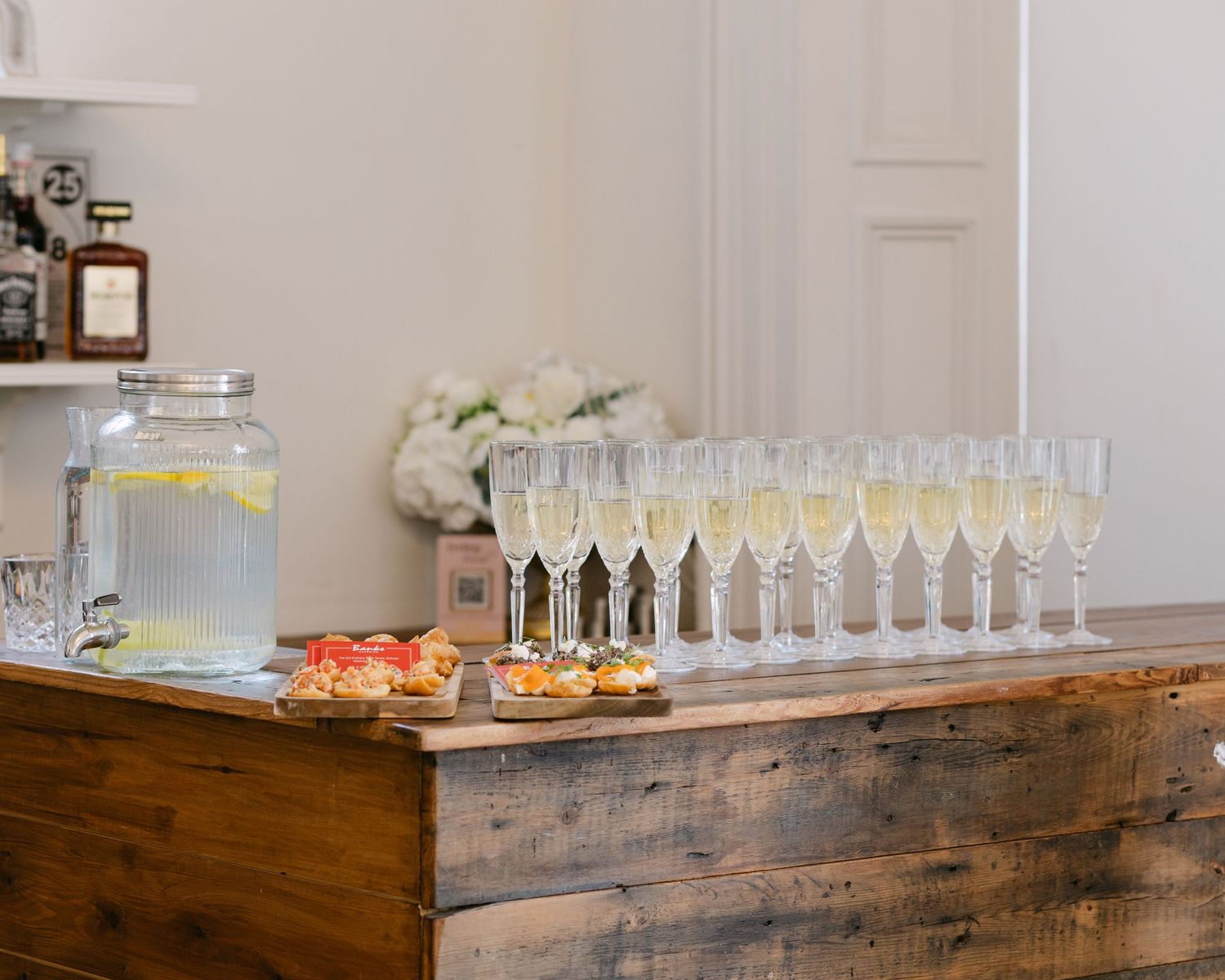 Champagne flutes and lemonade dispenser on rustic wooden bar with elegant appetizers at a wedding reception.