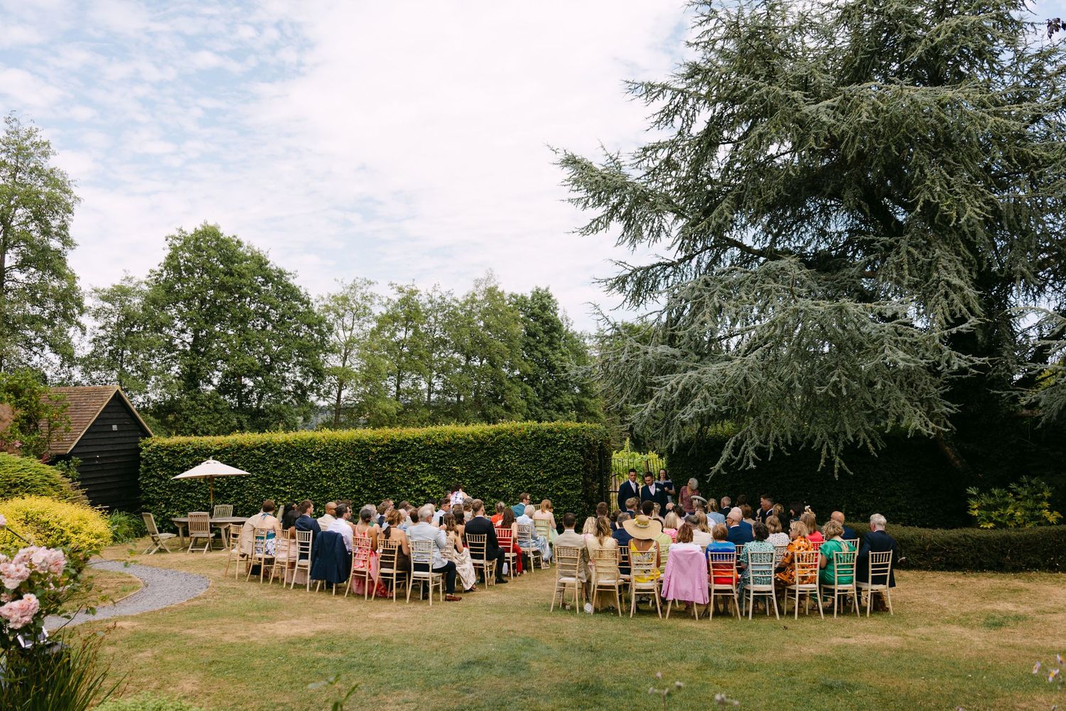 Outdoor wedding ceremony in a lush garden with guests seated at round tables beneath a blue sky.