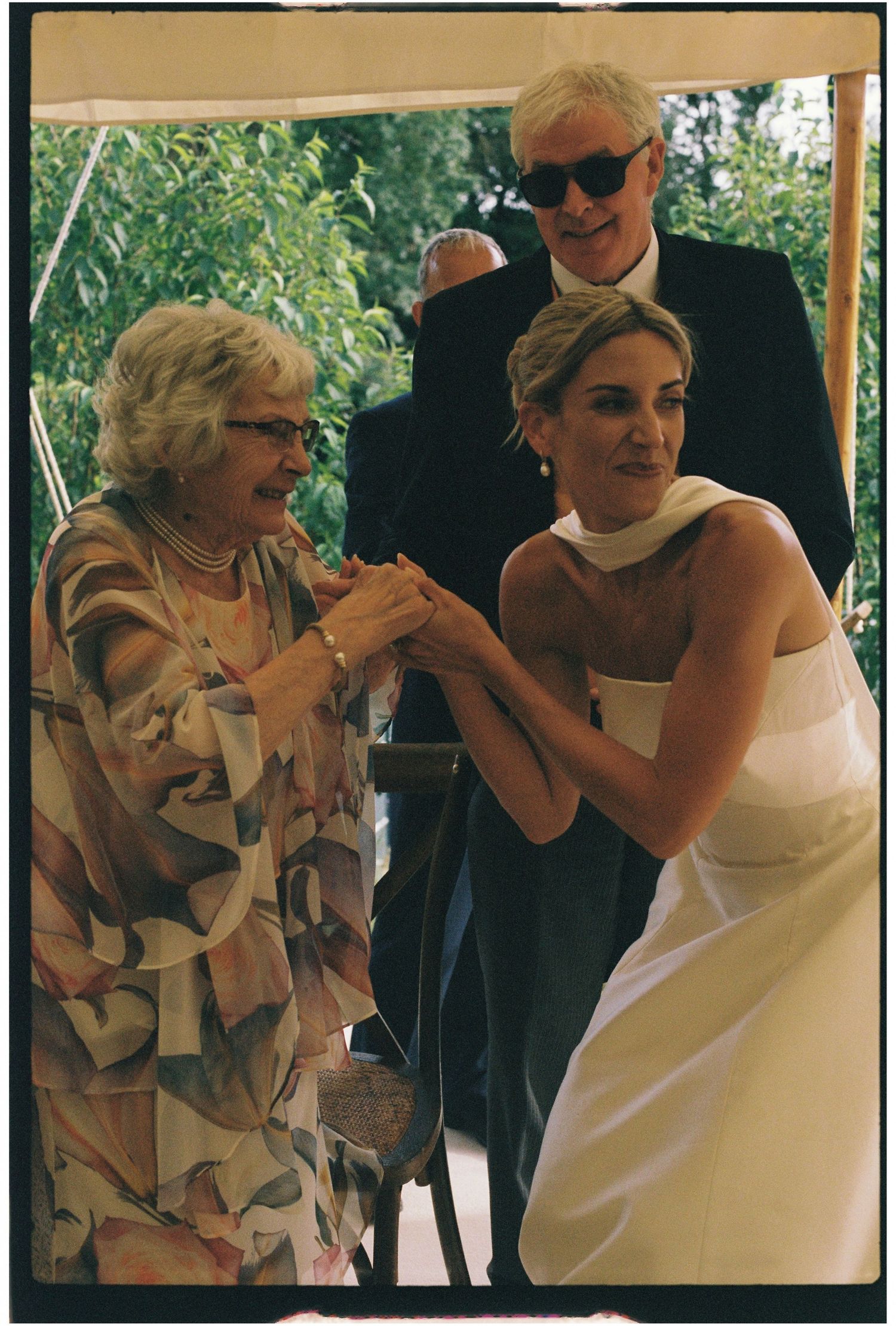 Bride in white gown holding hands with elderly woman at outdoor wedding ceremony.
