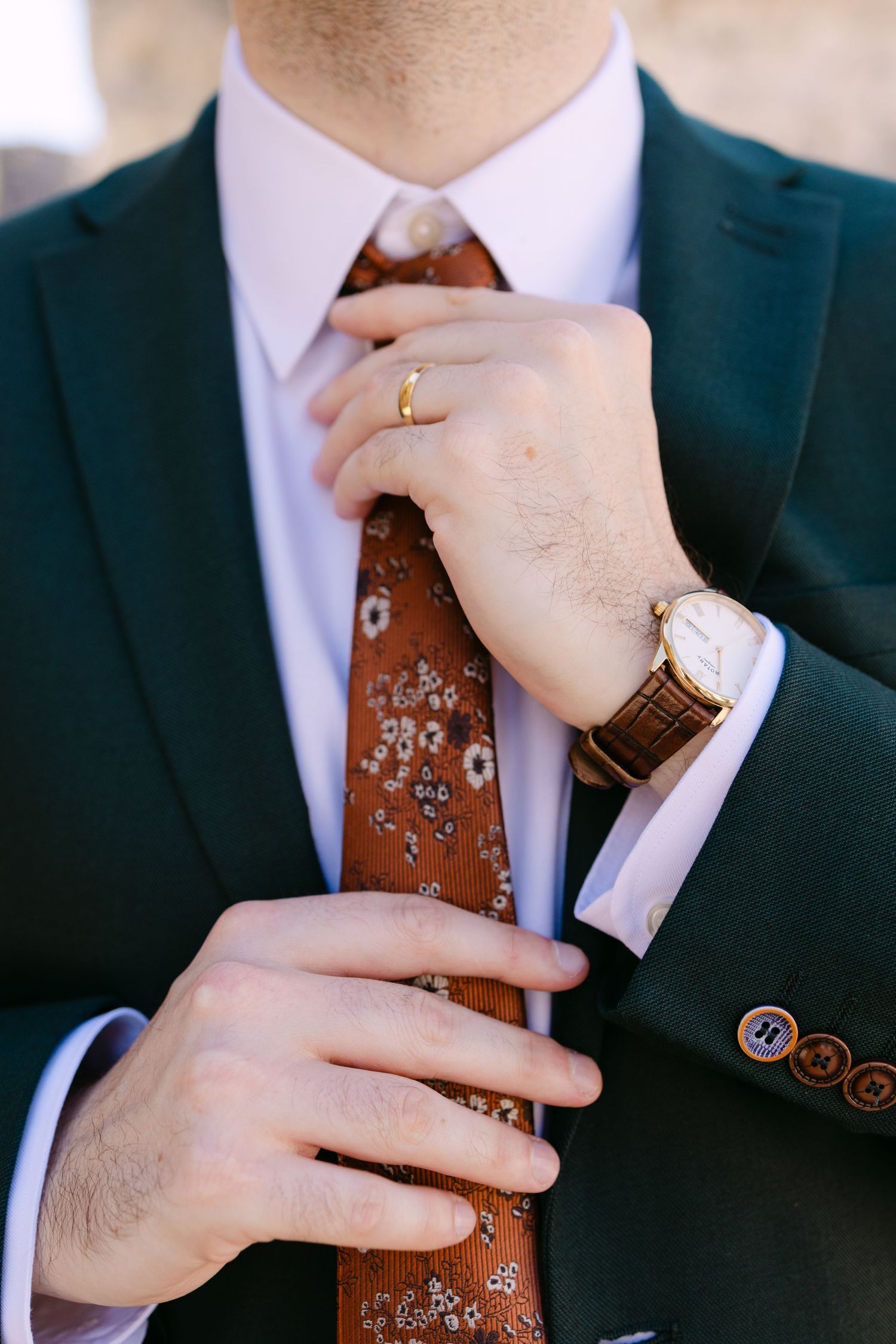 Man in green suit adjusting floral rust-colored tie, wearing gold watch and ring, close-up detail shot.