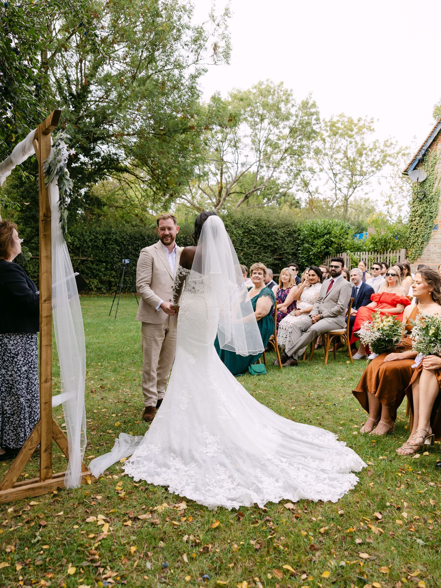 Outdoor wedding ceremony in a garden setting with a wooden arch draped in white fabric and guests seated on wooden chairs.