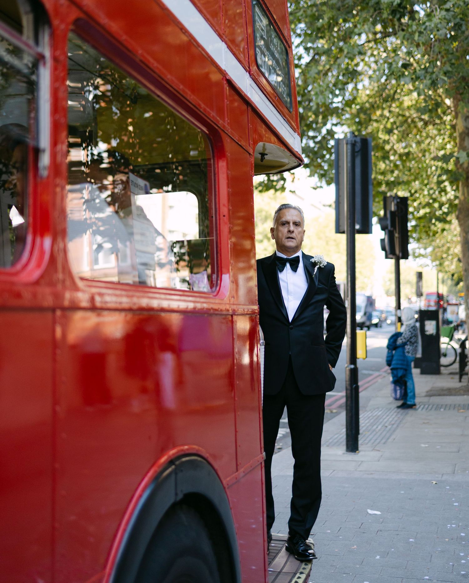 Person in formal black tuxedo standing next to vintage red London bus.