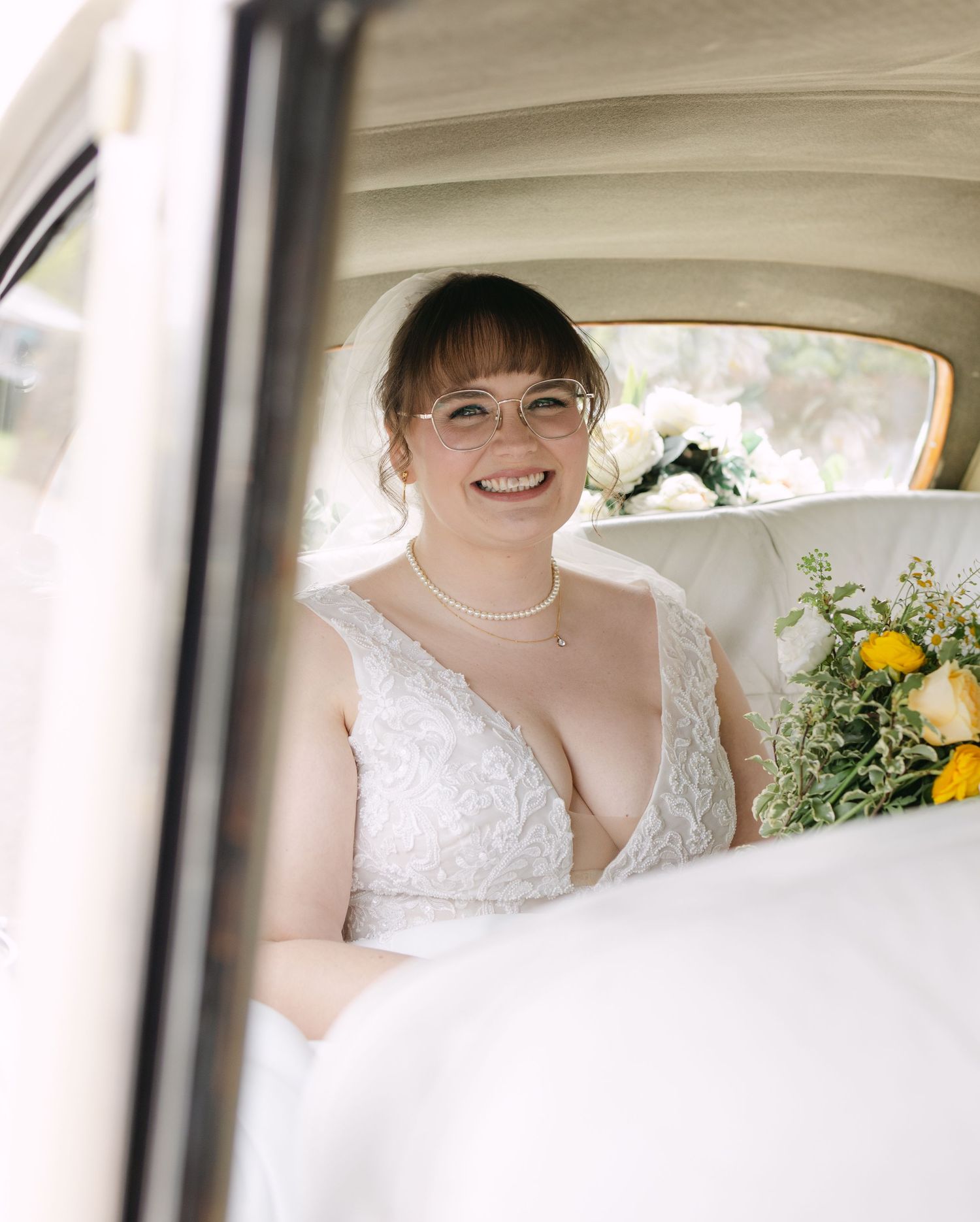 Smiling bride in white lace gown and pearl necklace holding yellow bouquet seated in vintage wedding car.