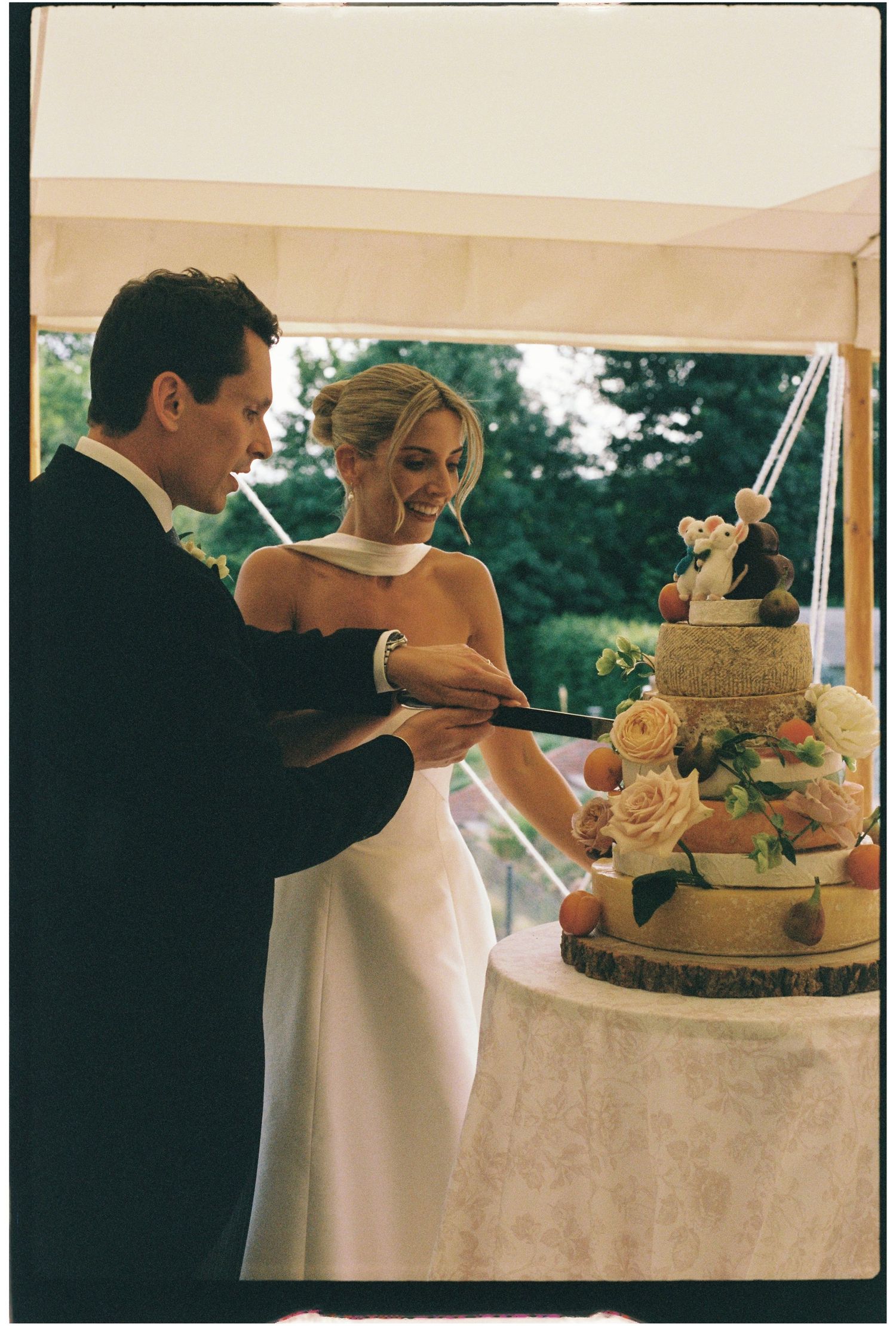 Couple cuts a tiered cheese wedding cake decorated with roses inside a marquee venue.