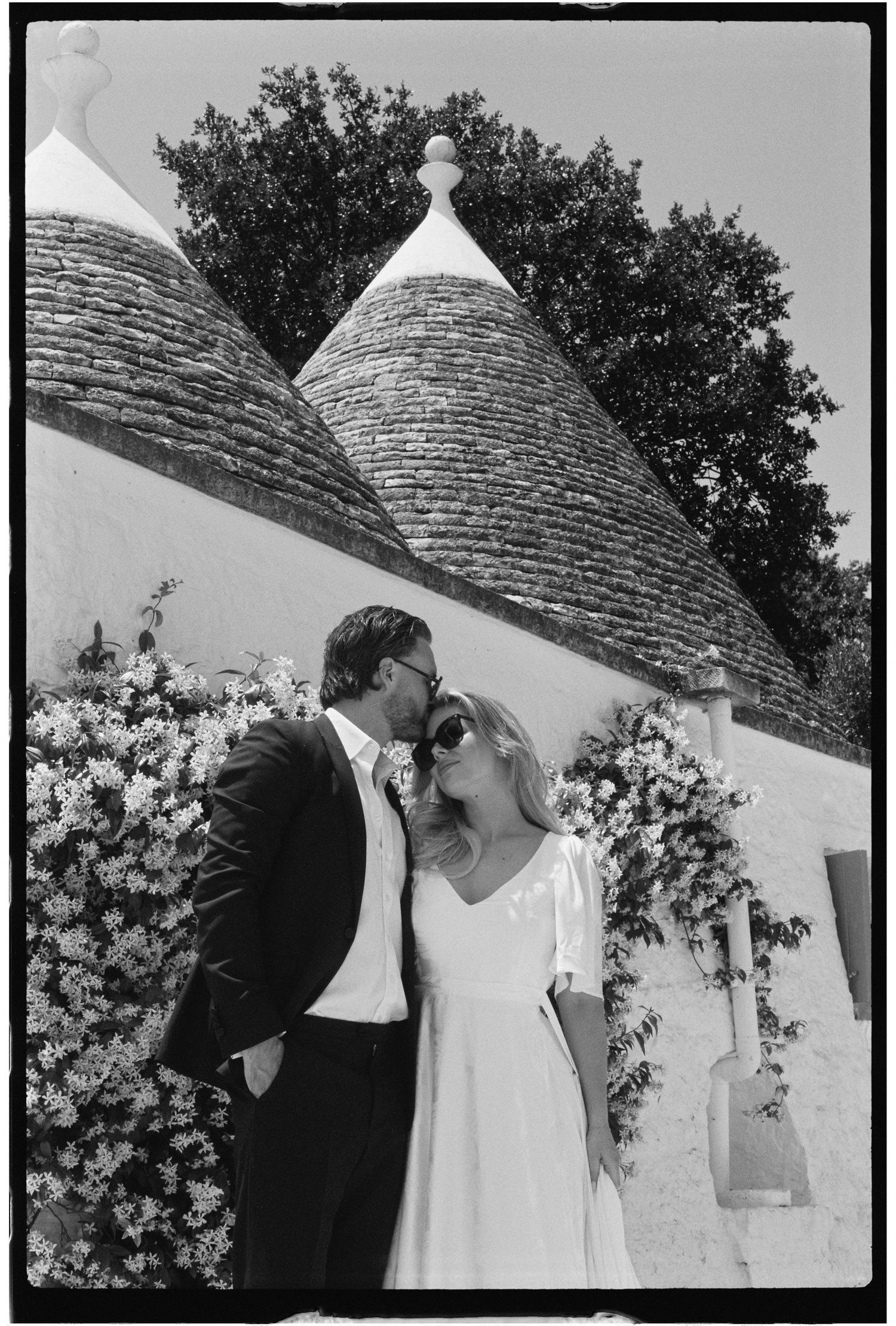 Black and white photo of a couple sharing a kiss near trulli stone buildings adorned with flowers.