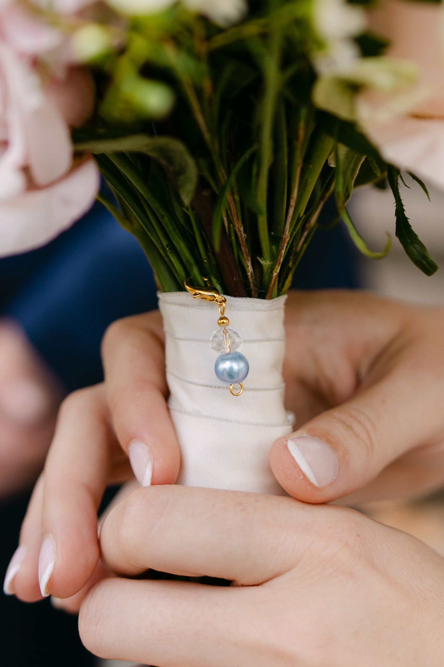 Hands holding a white wrapped bouquet stem decorated with a delicate blue pearl charm pendant.