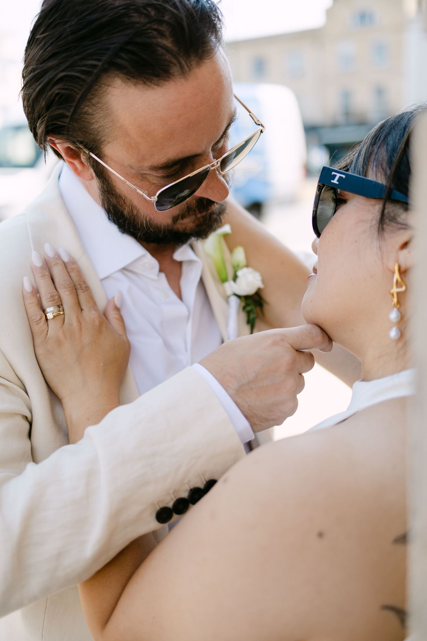 Groom in beige suit kissing bride's hand at outdoor wedding ceremony, both wearing sunglasses and gold rings.