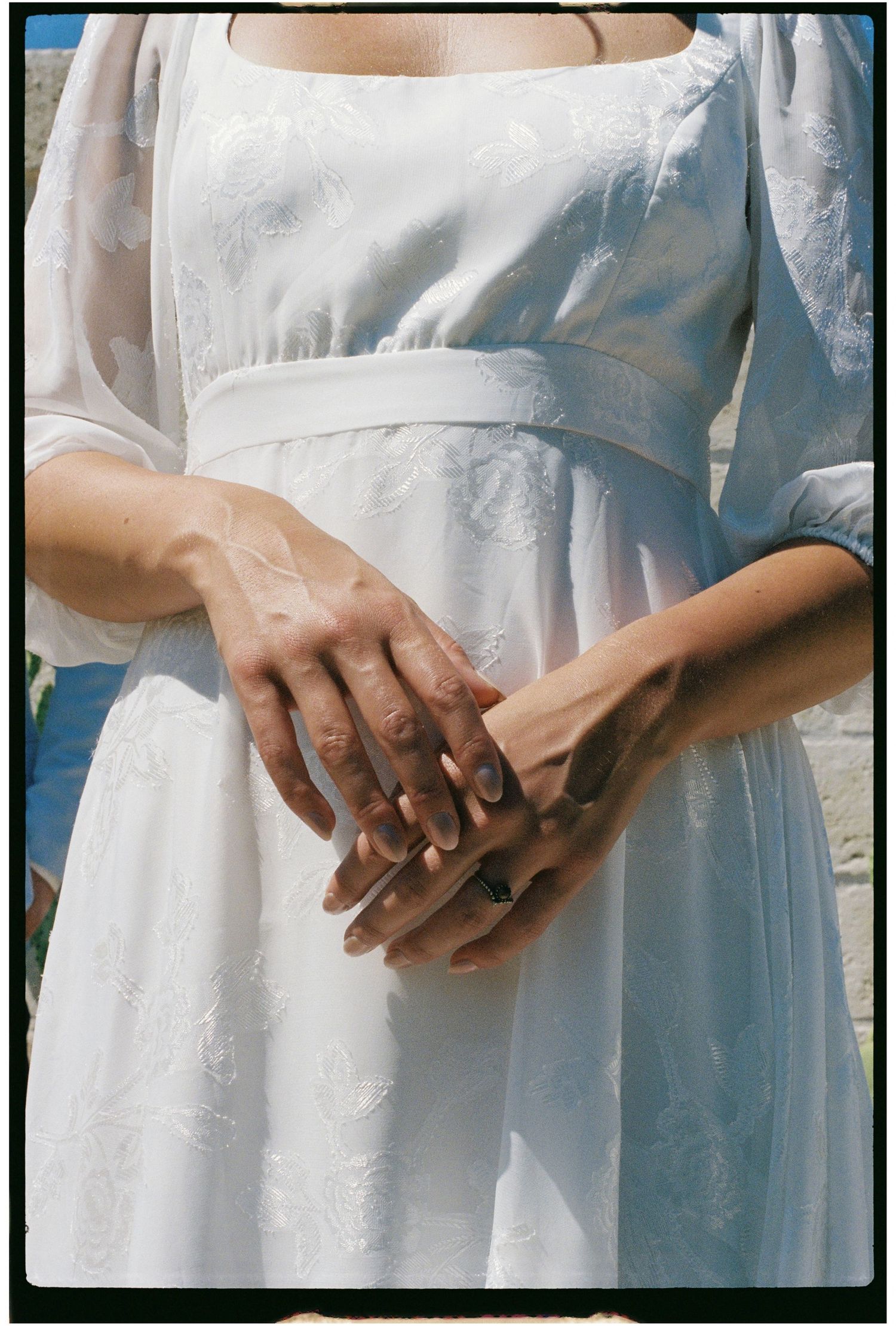 Close-up of couple's hands clasped together, bride wearing white embroidered dress and delicate ring.