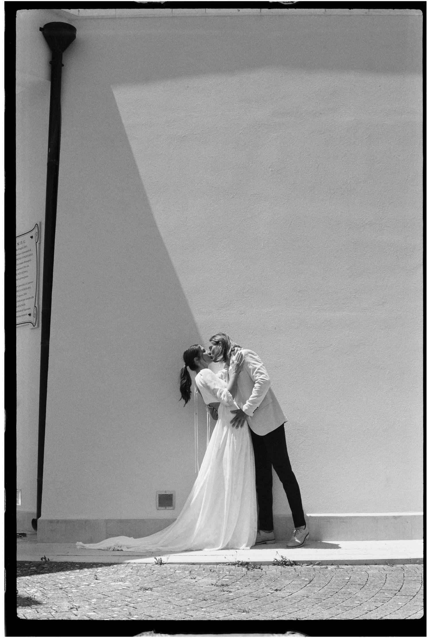 Black and white wedding photo of couple kissing against a sunlit white wall outdoors.