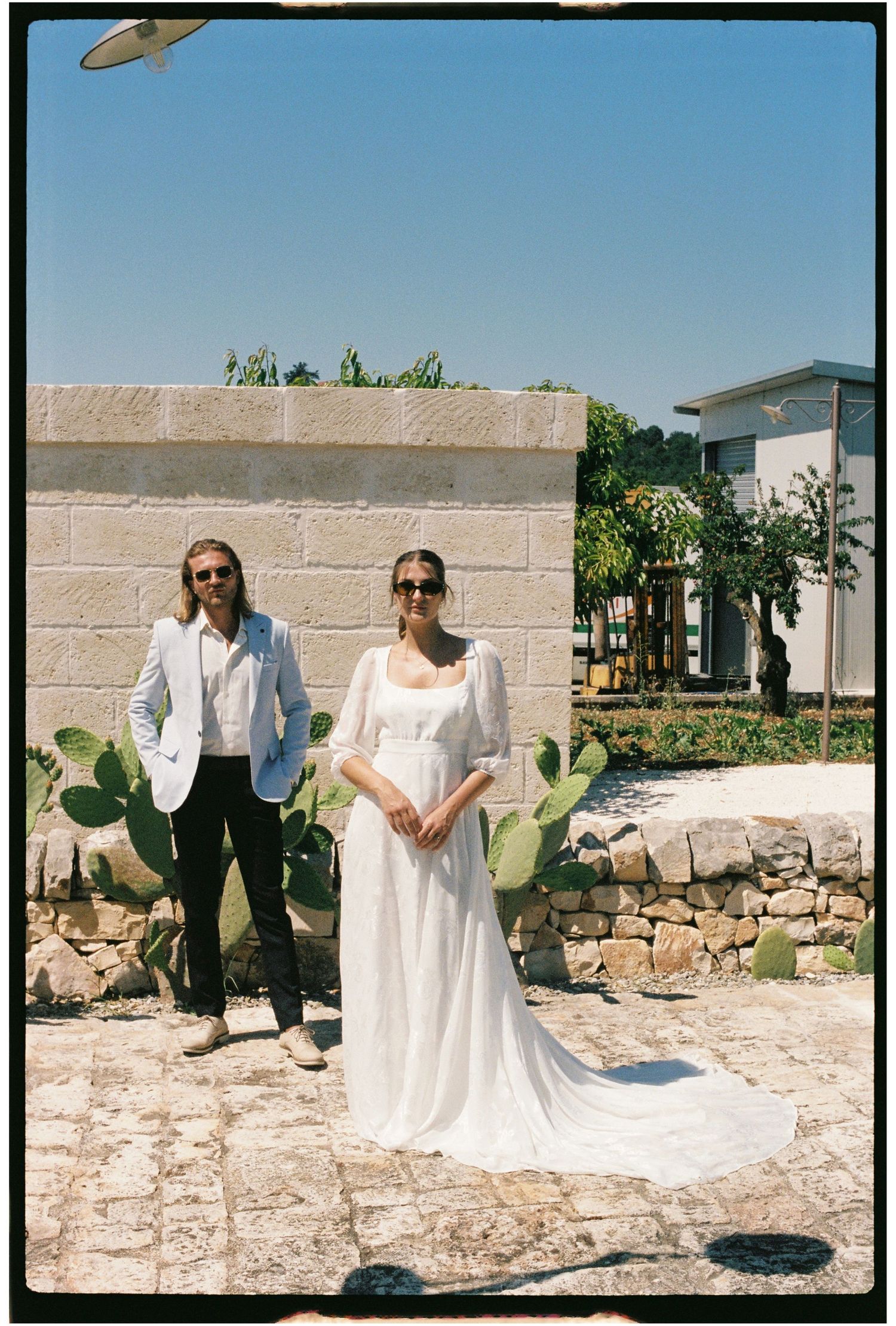 Bride in flowing white gown and groom in light blazer pose outdoors at a sunny Mediterranean wedding venue.