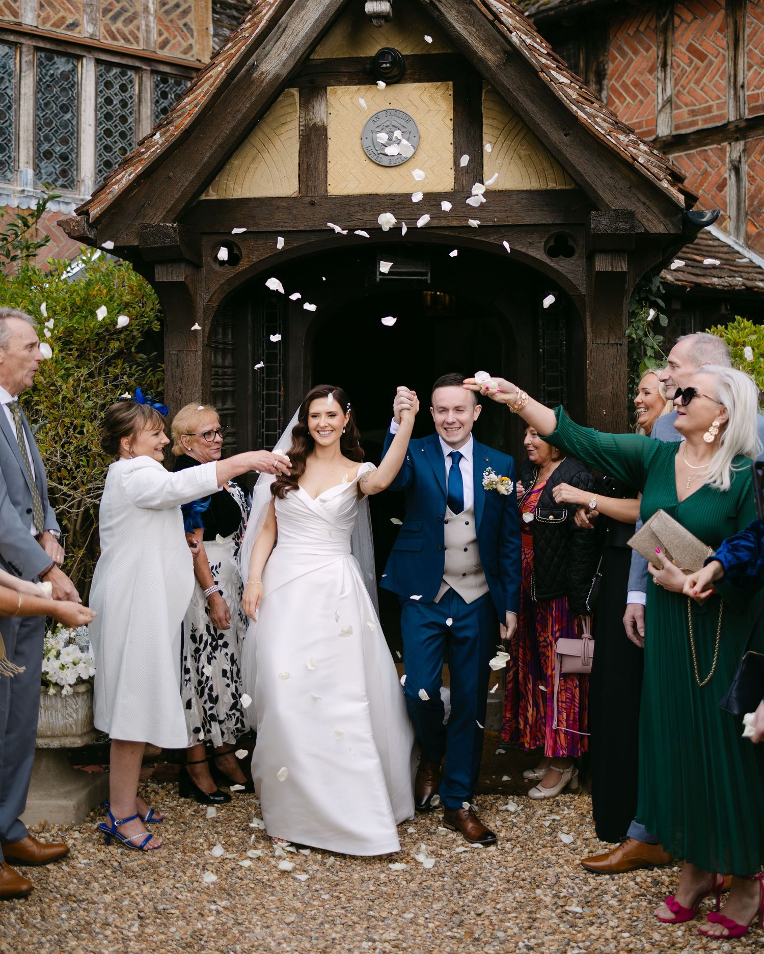Wedding party celebrates with confetti outside a Tudor-style church entrance.