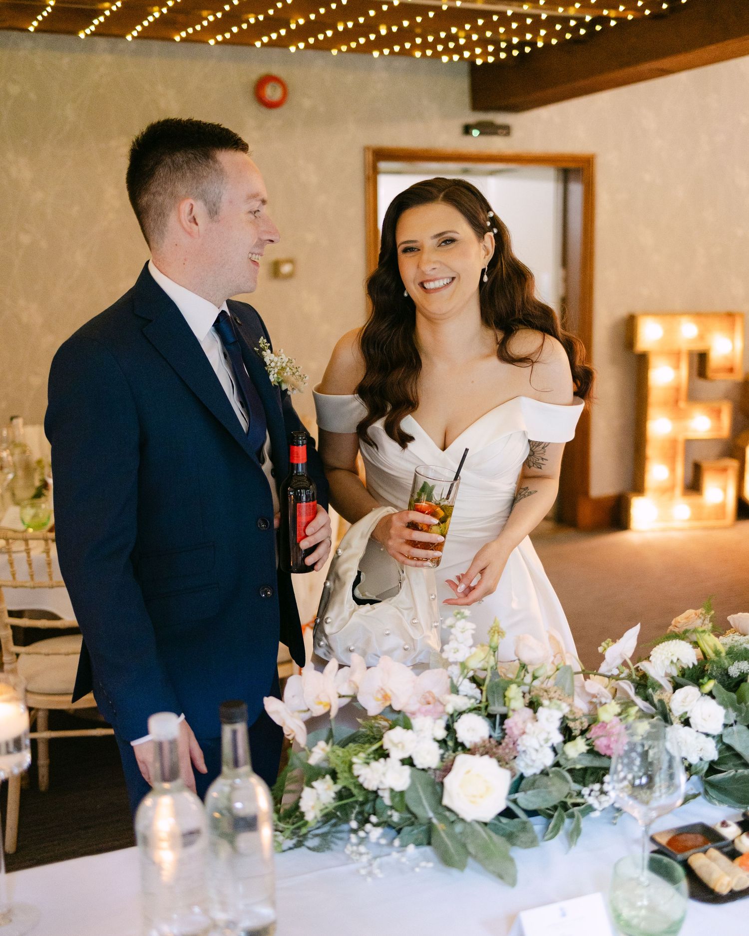 Newlyweds share drinks at their reception head table decorated with white flowers.