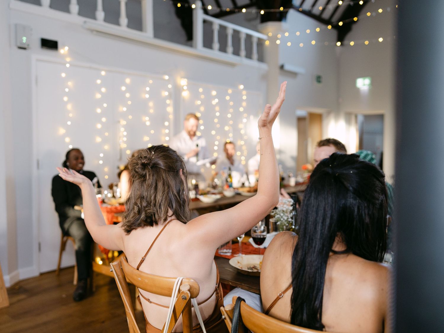Wedding guests celebrating with raised arms under string lights at an indoor reception.