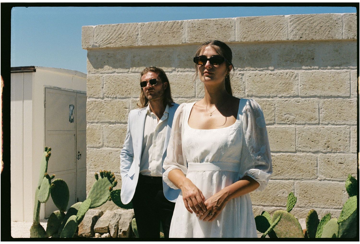 Couple posing outdoors in Puglia, woman in white dress and man in light suit, with cactus plants.