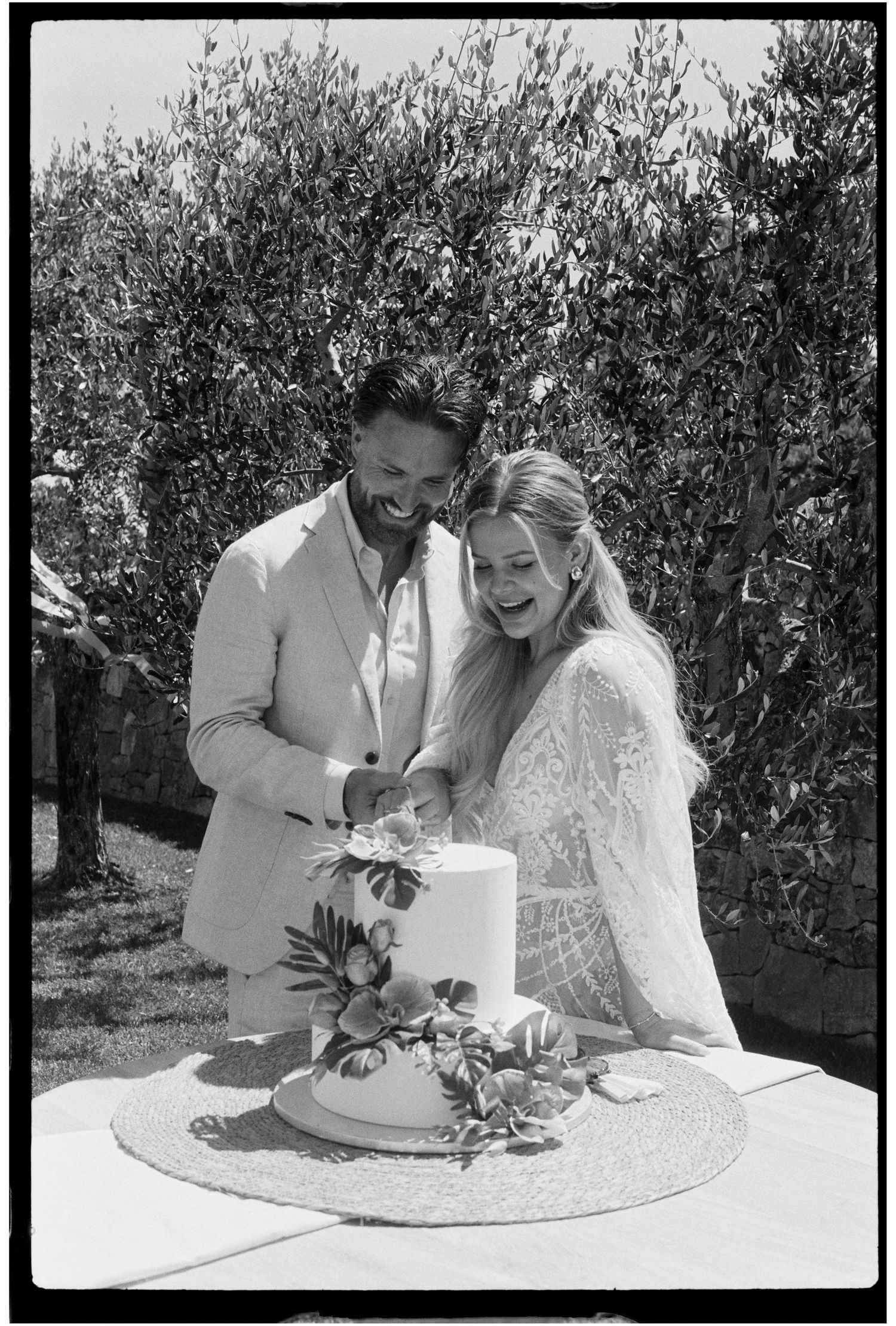 Laughing couple cuts their wedding cake outdoors surrounded by lush greenery.