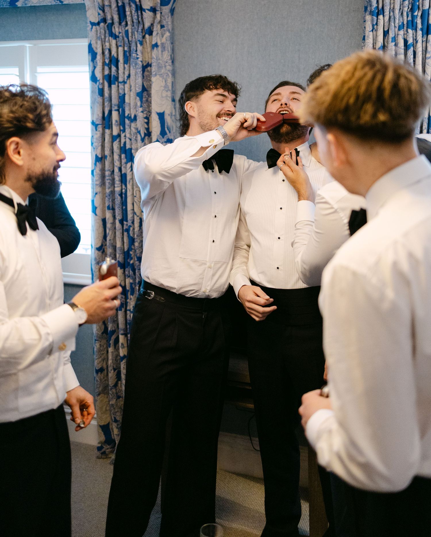Groomsmen adjusting their bow ties and getting ready in a blue-curtained room.