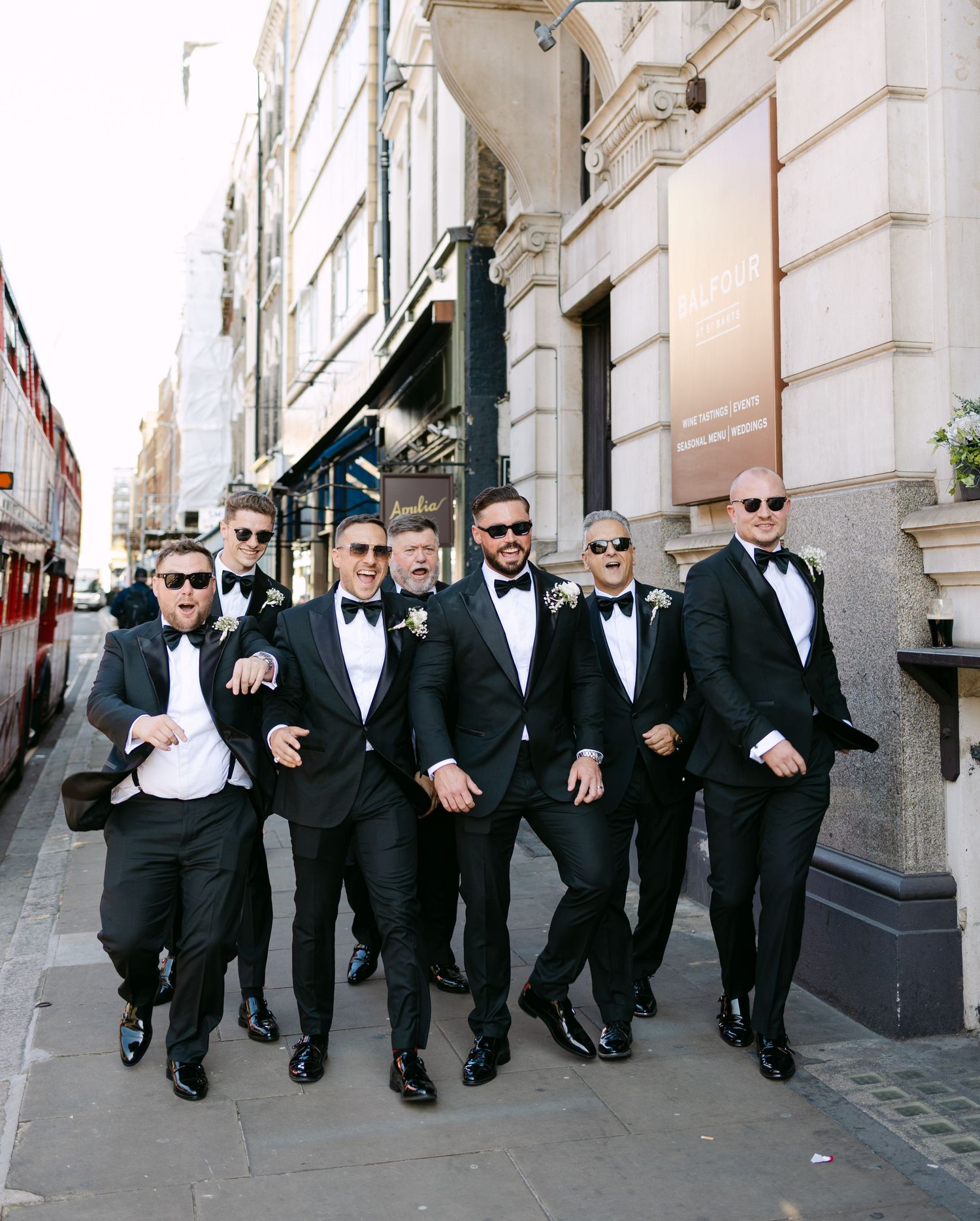 Groomsmen in black suits walk together down urban sidewalk during wedding celebration.