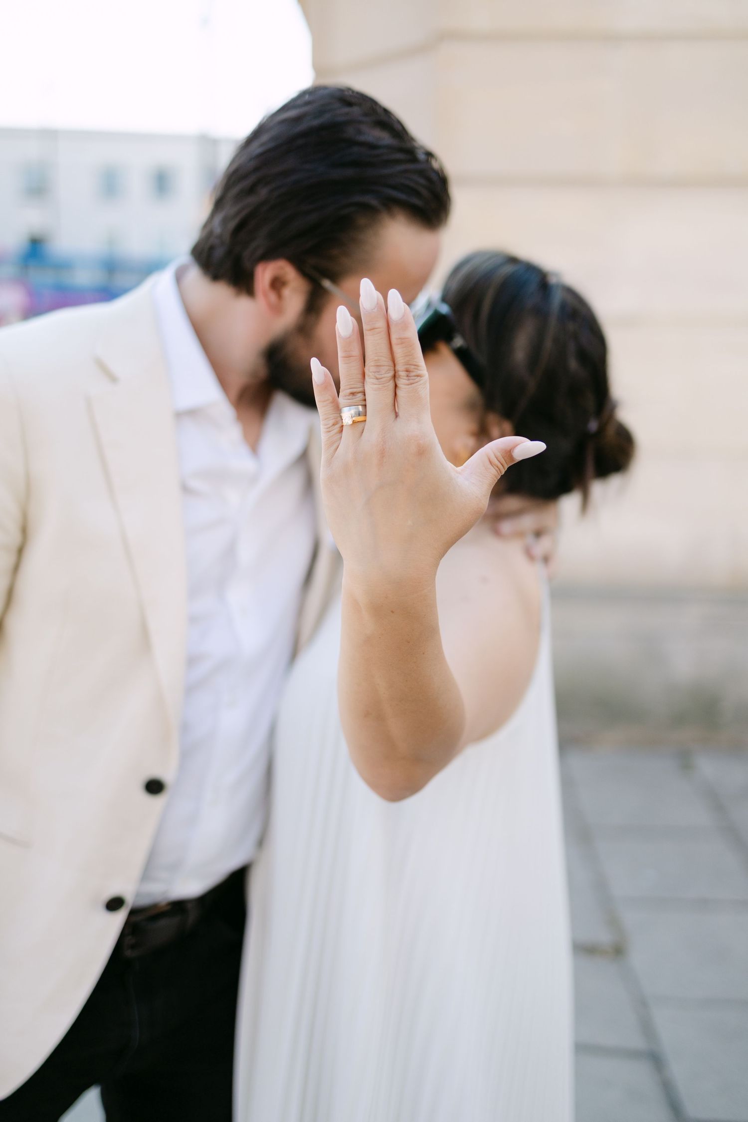 Bride shows off engagement ring while groom kisses her cheek, both dressed in white and beige wedding attire outdoors.