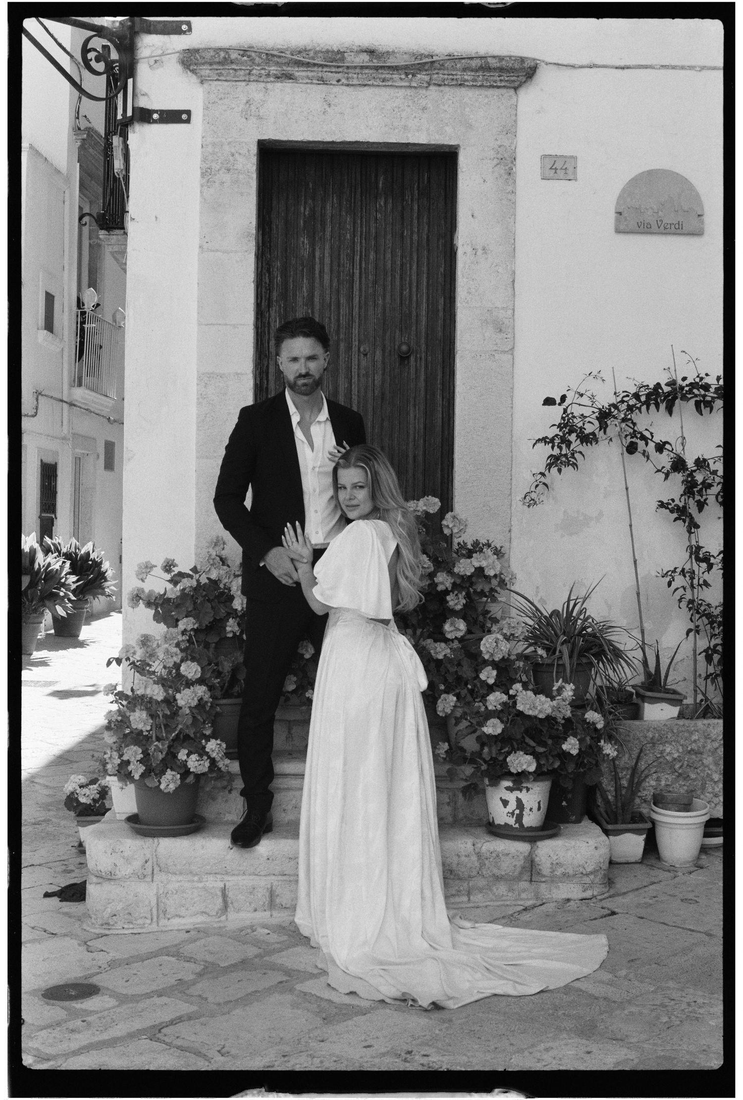 Bride in elegant white gown and groom in dark suit posing before a rustic stone doorway with blooming flowers.