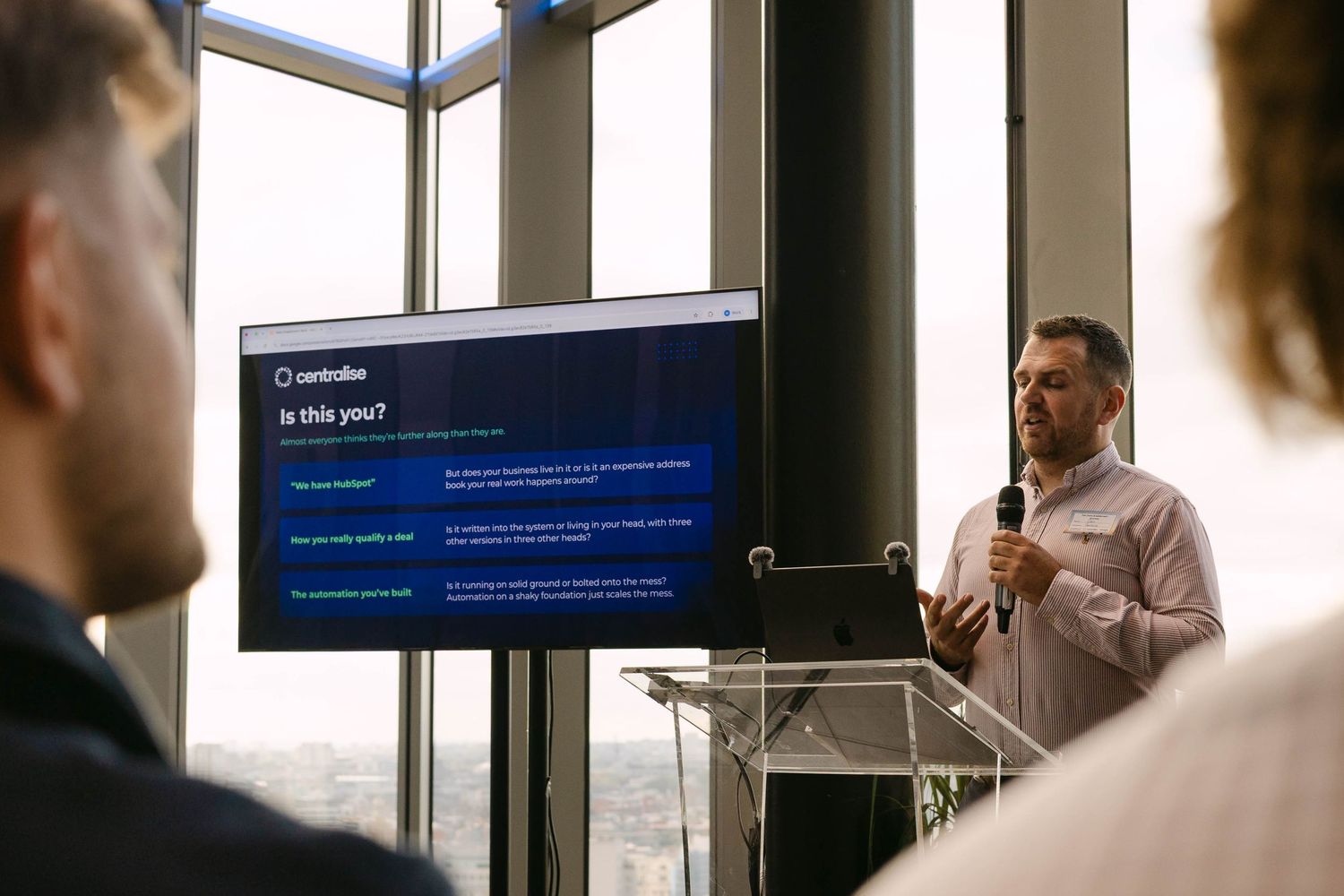 Business presenter speaks at podium beside a screen displaying a Controller software presentation in a modern office.