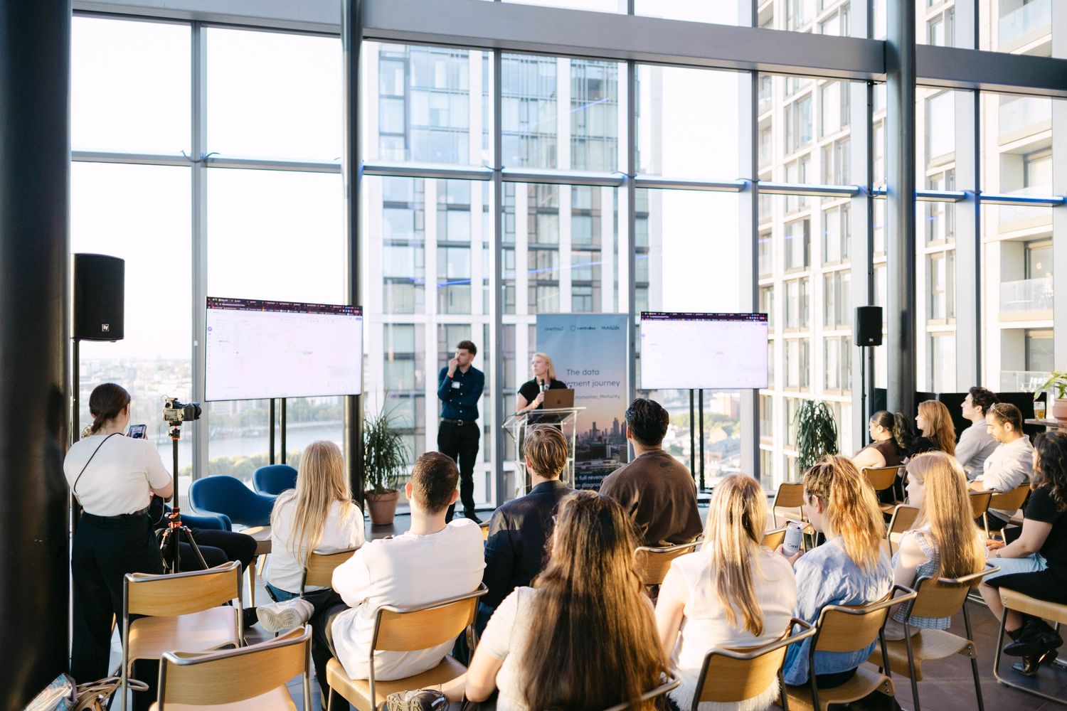 Professionals attend a business conference in a modern glass-walled venue with a speaker presenting at the podium.