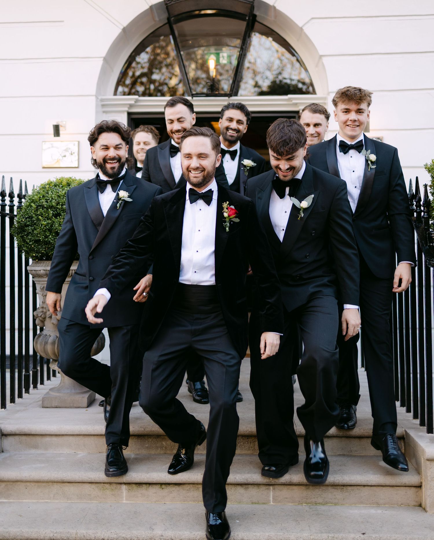 Groomsmen in black tuxedos pose on stone steps with architectural arch in background.