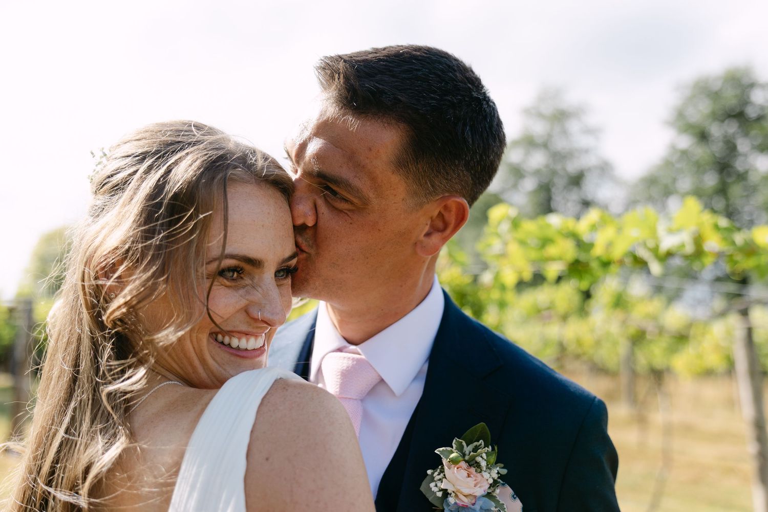 A groom in a navy suit kisses his smiling bride on the cheek in a vineyard on their wedding day.