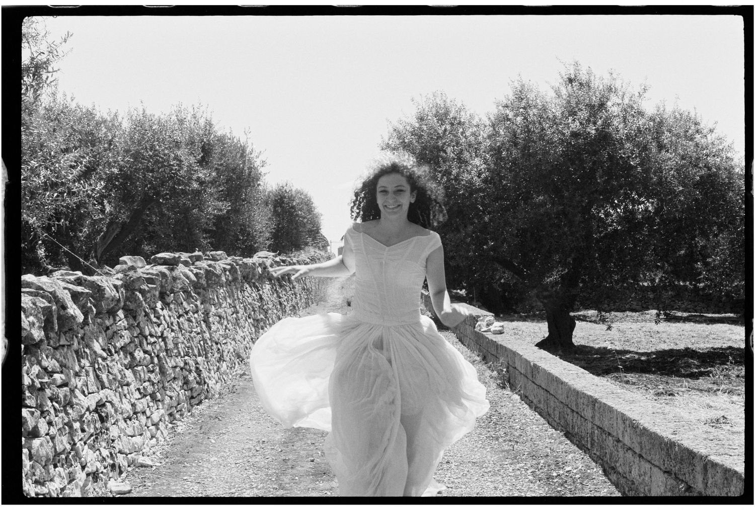 A woman in a flowing white dress runs along a stone-walled path lined with olive trees in a black and white photo.